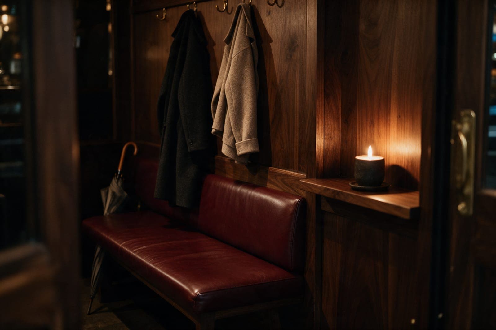 An oxblood leather bench against a dark paneled wall, two wool coats on brass hooks above it, a candle on a ledge.