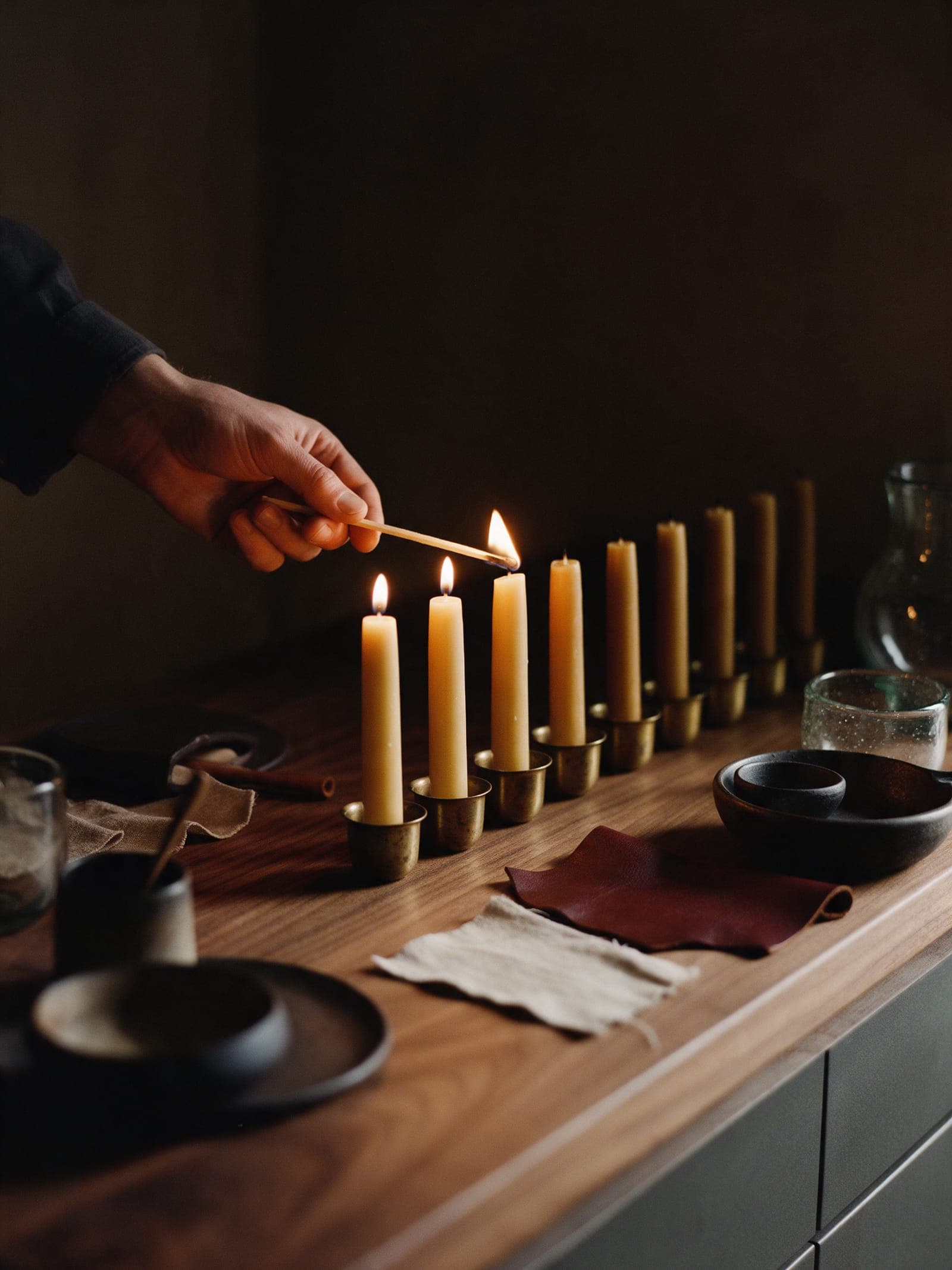 A hand with a long match lighting beeswax candles along the walnut counter before service, the first three lit.
