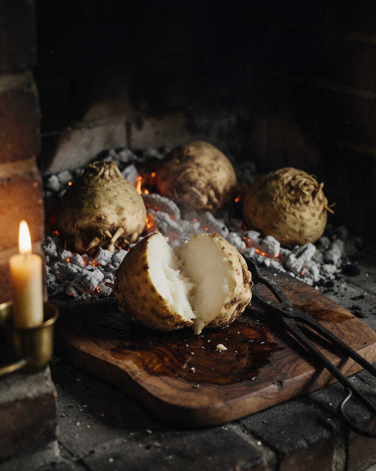 Whole celeriac roots baking in the hearth's ash and embers, one split open to show dense ivory flesh, iron tongs beside it.
