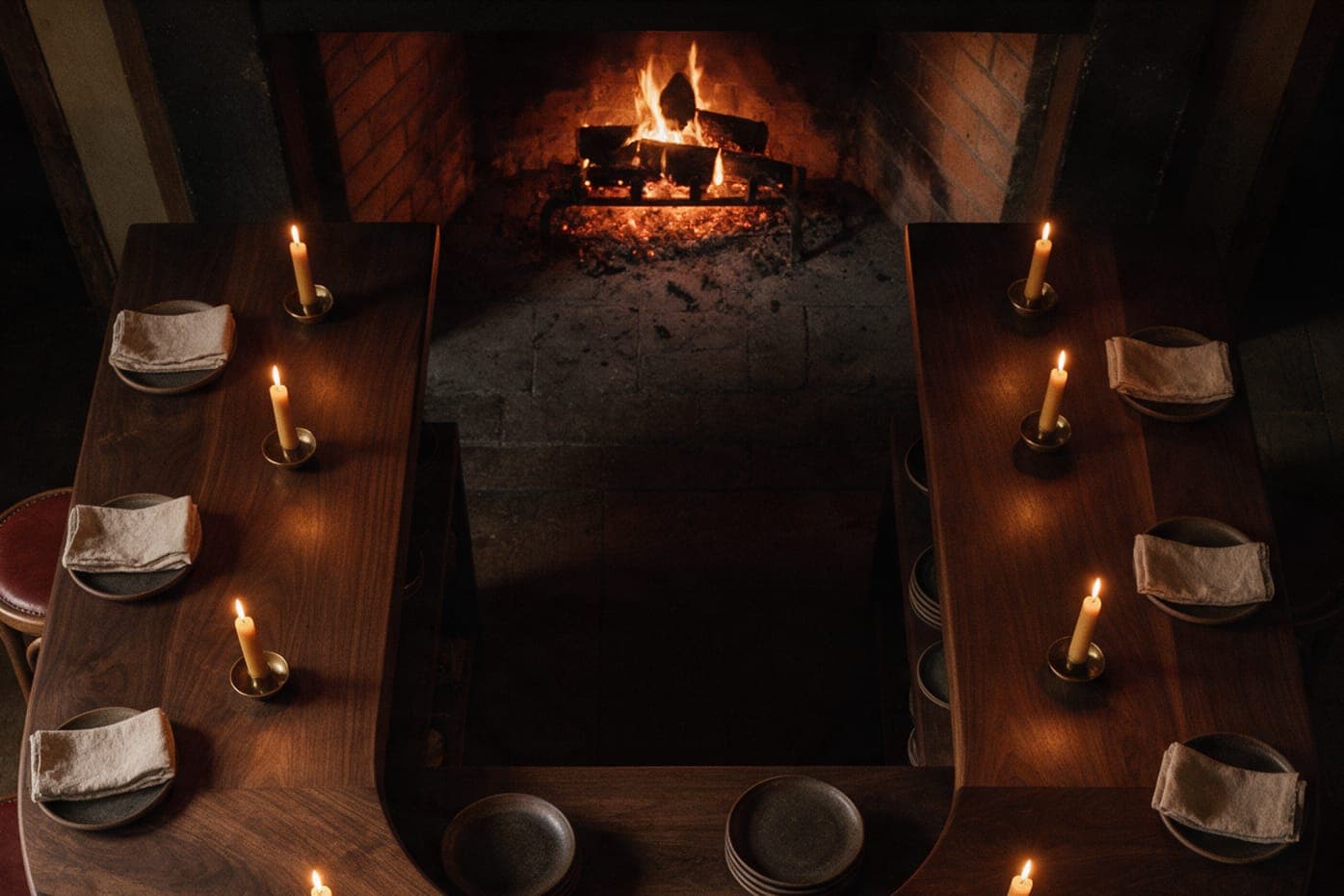 The hearth end of the horseshoe from above: both walnut arms set with linen, dark stoneware and a lit candle at each place, the fire glowing between them.