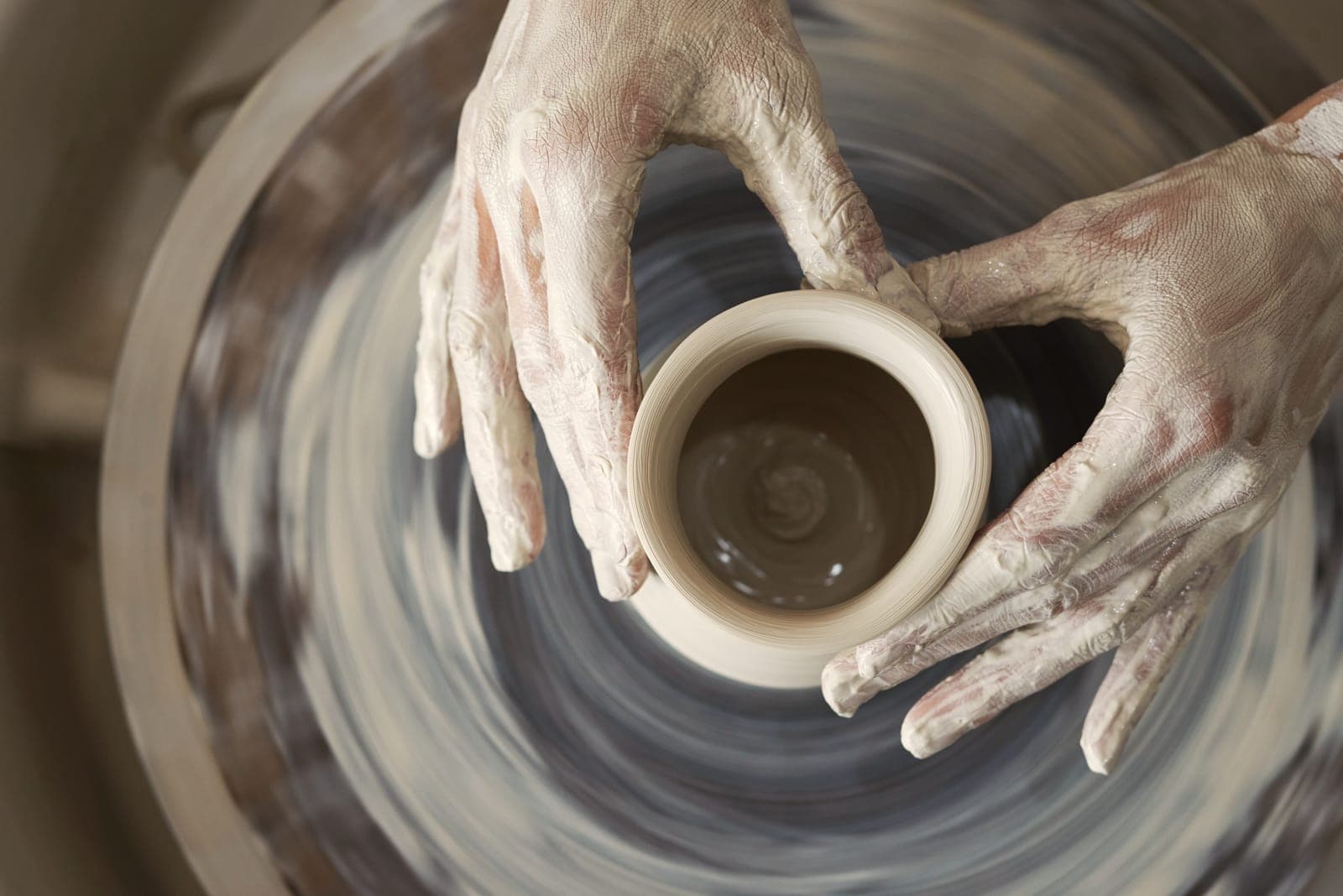 From above: slip-covered hands shaping a small cup on the spinning wheel