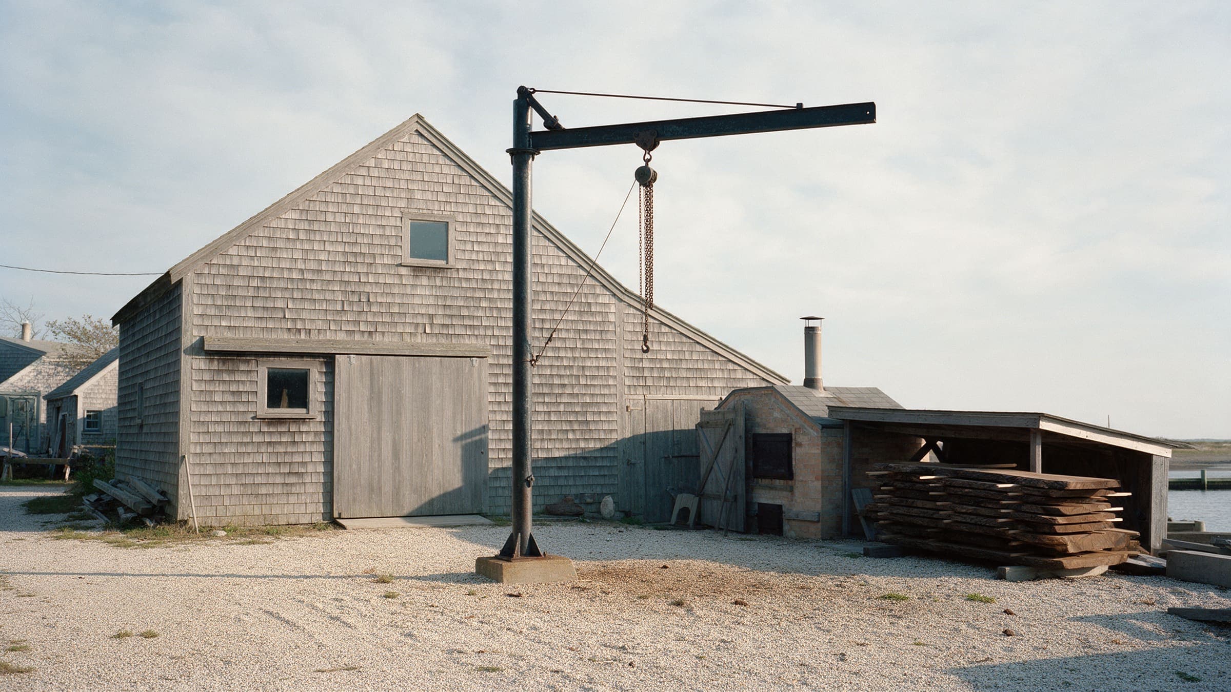 The cedar-shingled boat shop with the old jib crane in the gravel yard, the kiln shed and stacked walnut slabs by the harbor
