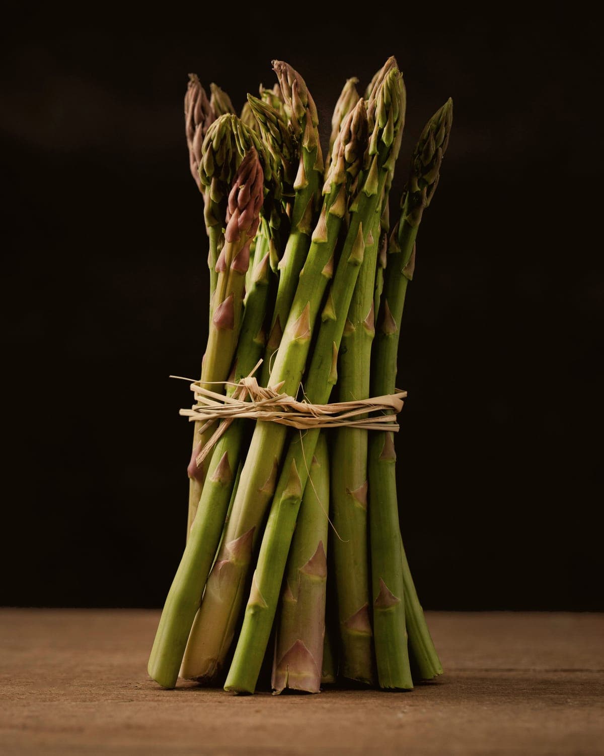 A bundle of green asparagus tied with raffia, standing on a wooden table against black.
