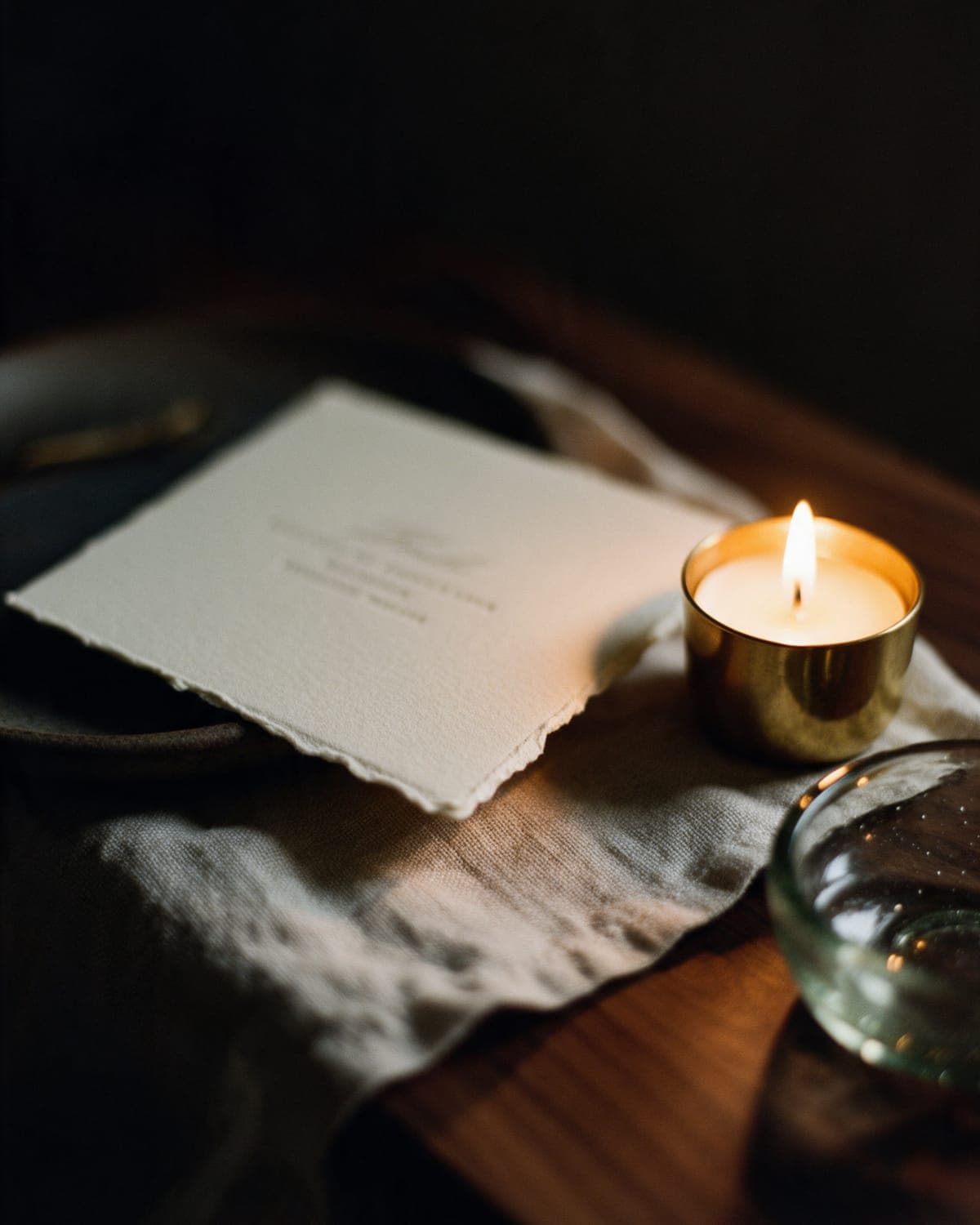 A deckled cream menu card on undyed linen beside a brass candle cup and a water glass.