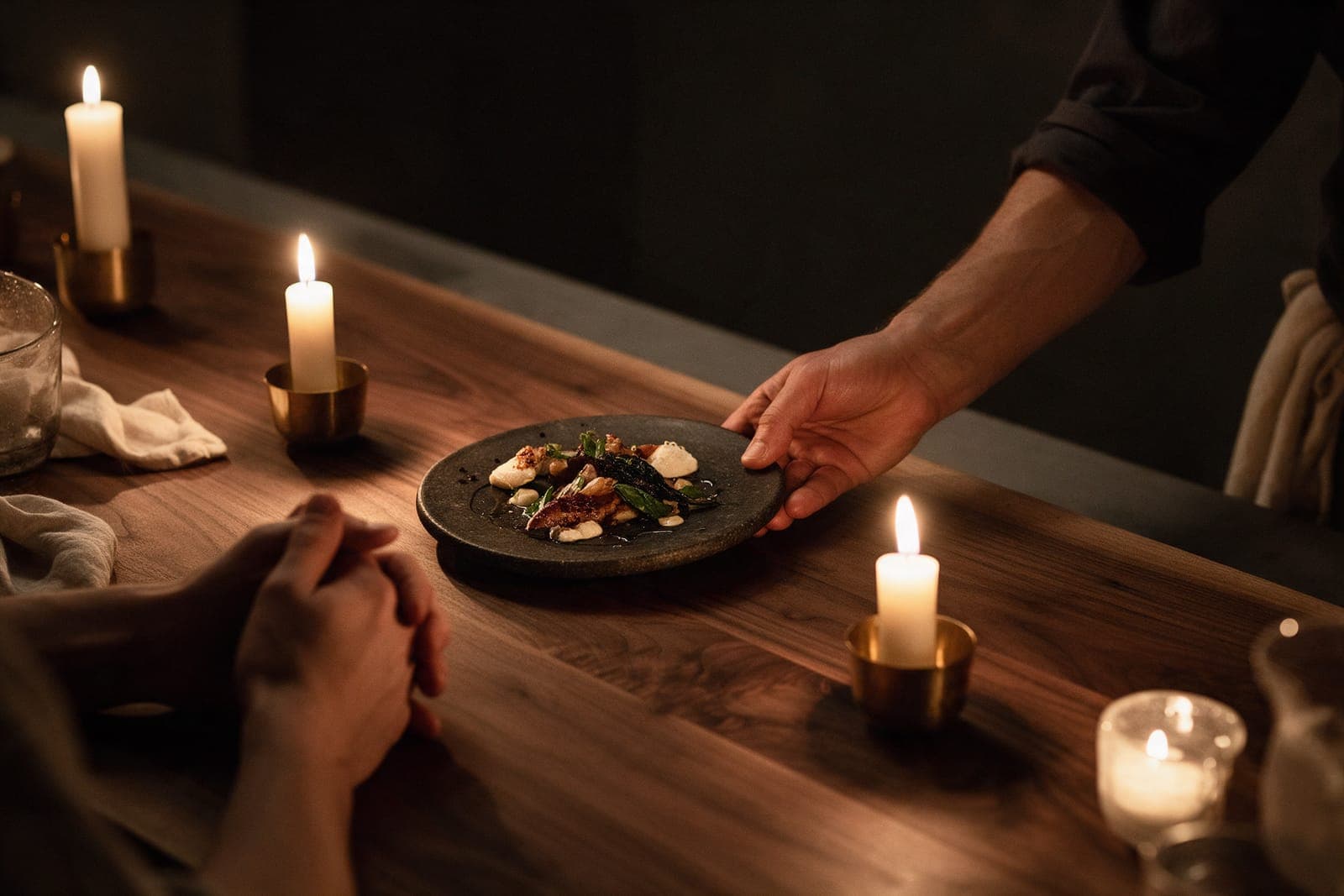 A cook's sleeved arm setting a dark plate down on the walnut counter in front of a guest's folded hands.
