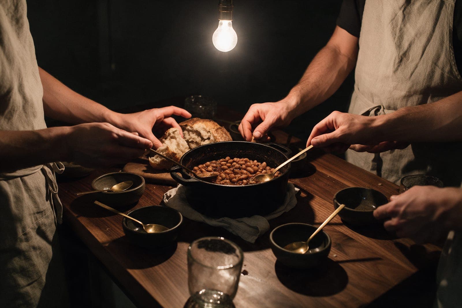 Hands and aproned forearms around a cast-iron pot of beans and torn bread on the walnut counter before service.