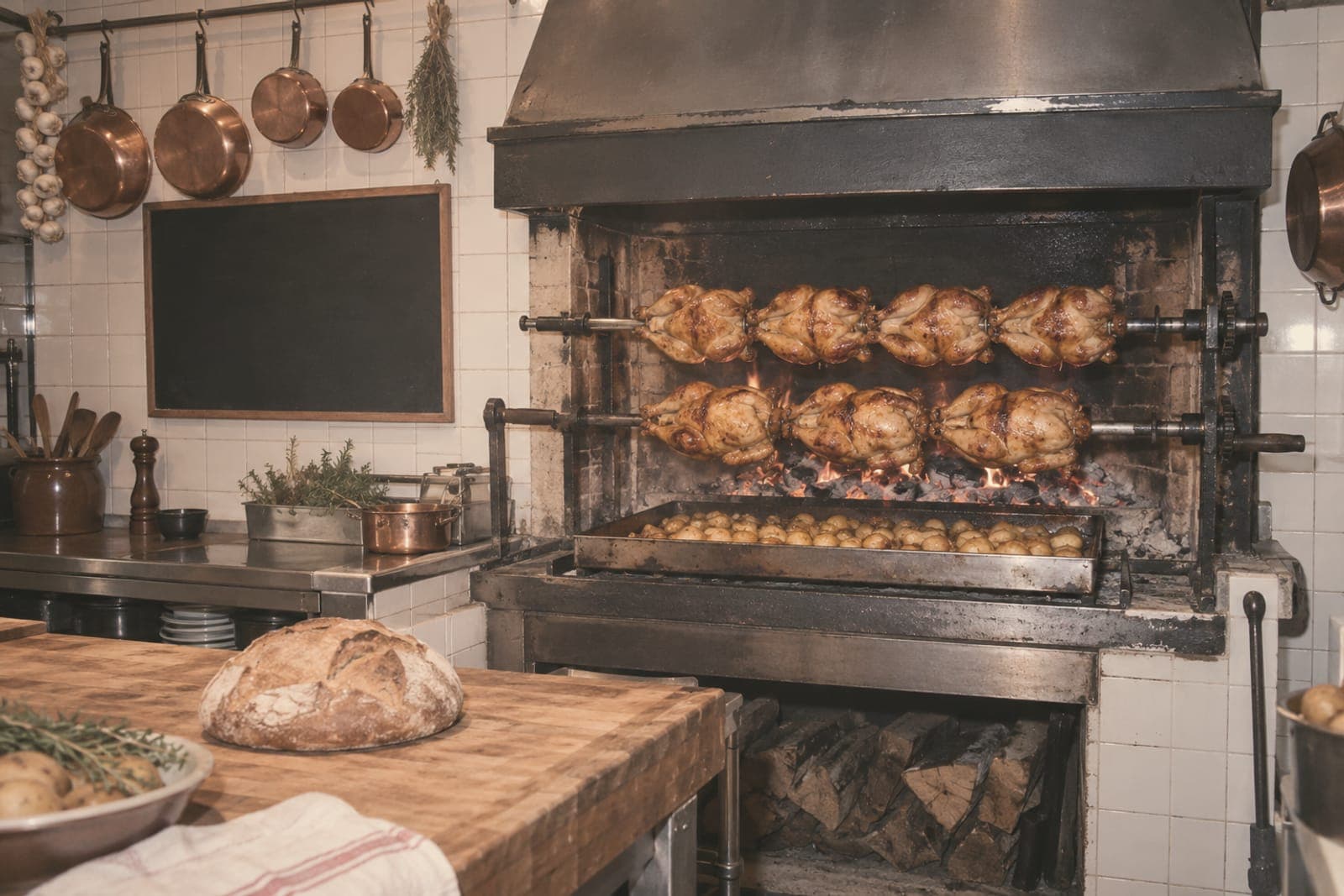 A faded color snapshot of a tiled kitchen: chickens turning on a wall rotisserie over embers, a tray of potatoes beneath, copper pans, a round loaf on the block.