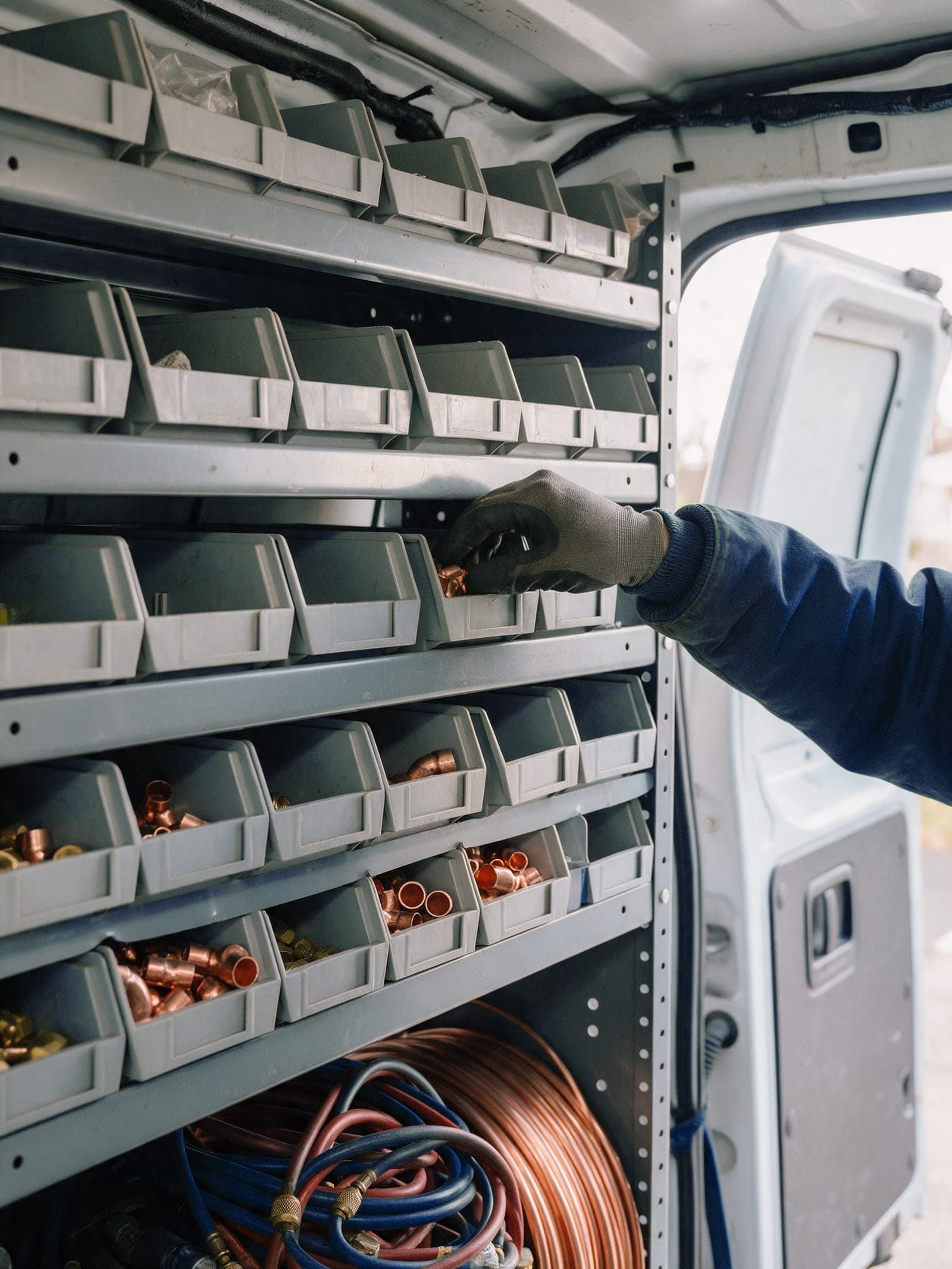 Gloved hand reaching into a parts bin on the steel shelving inside a service van