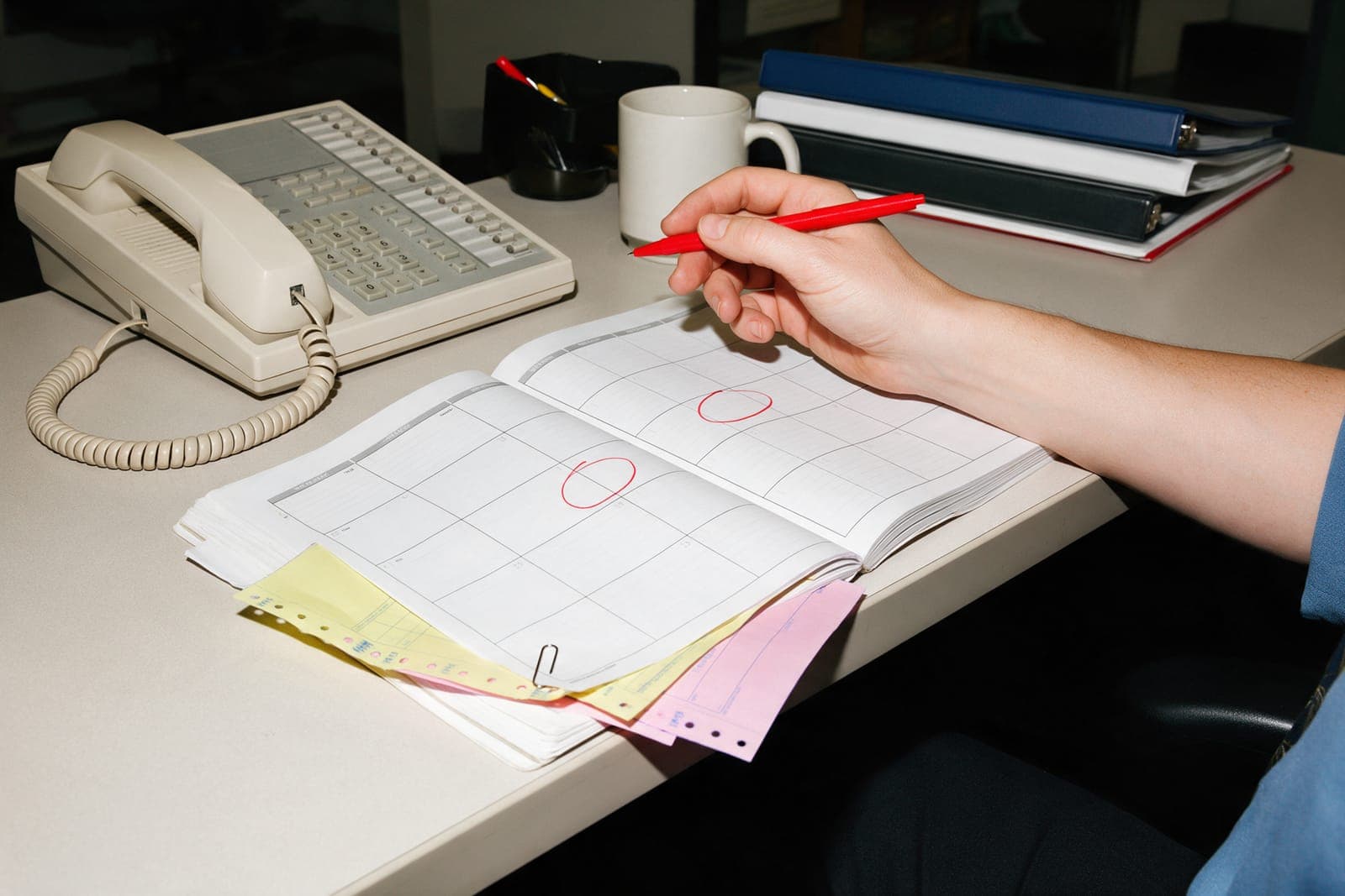 An office desk with a beige phone and a paper planner, two days circled in red, carbon copies clipped to its edge.