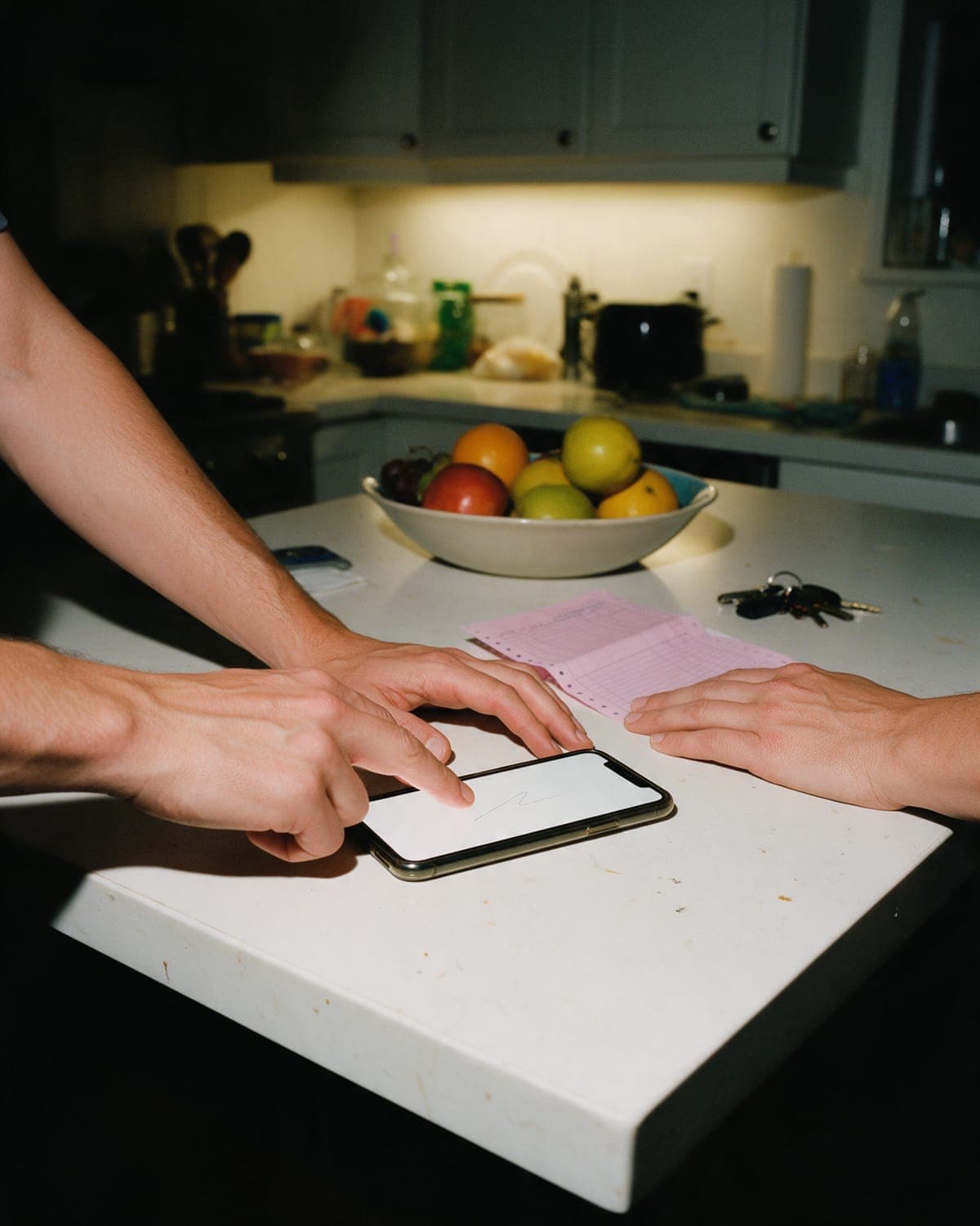 A finger signing on a phone lying on a kitchen island at night, a pink carbon copy and a second person's hand beside it.