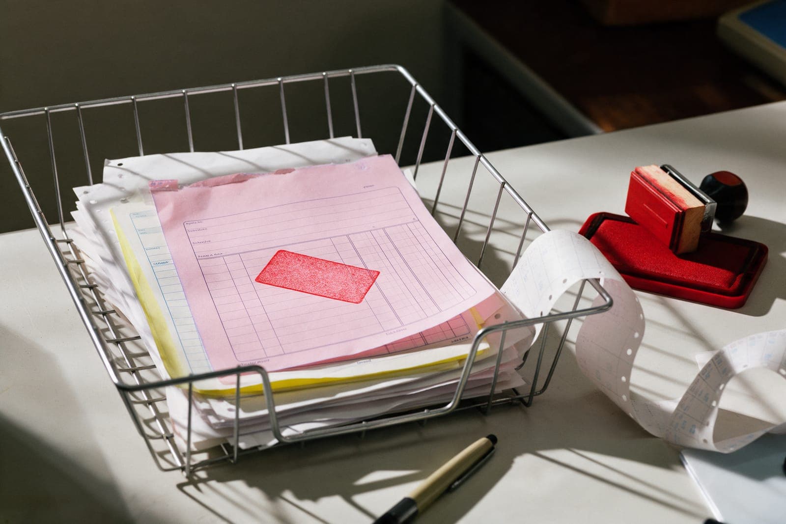 A wire inbox tray of invoices with a pink copy marked by a red rubber stamp, the stamp and ink pad beside it.