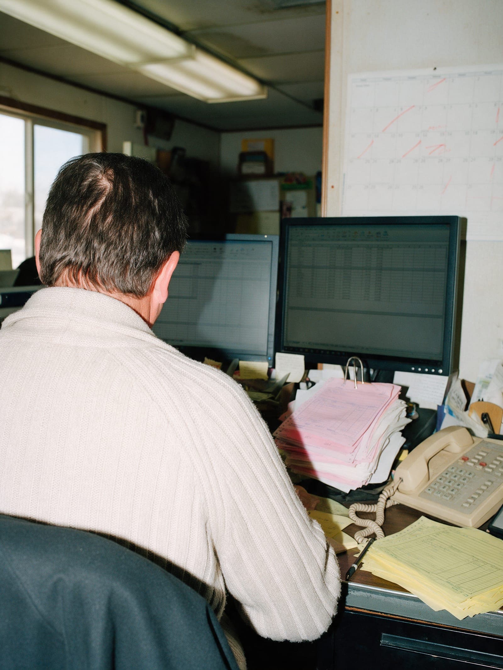 An office manager at a desk seen from behind, two monitors and a spike file of pink copies.