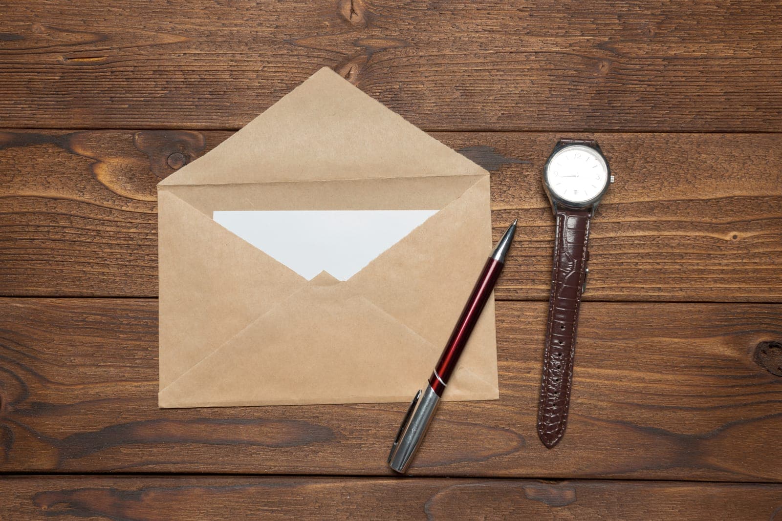An open kraft envelope with a blank card, a pen and a wristwatch on a dark wooden table.
