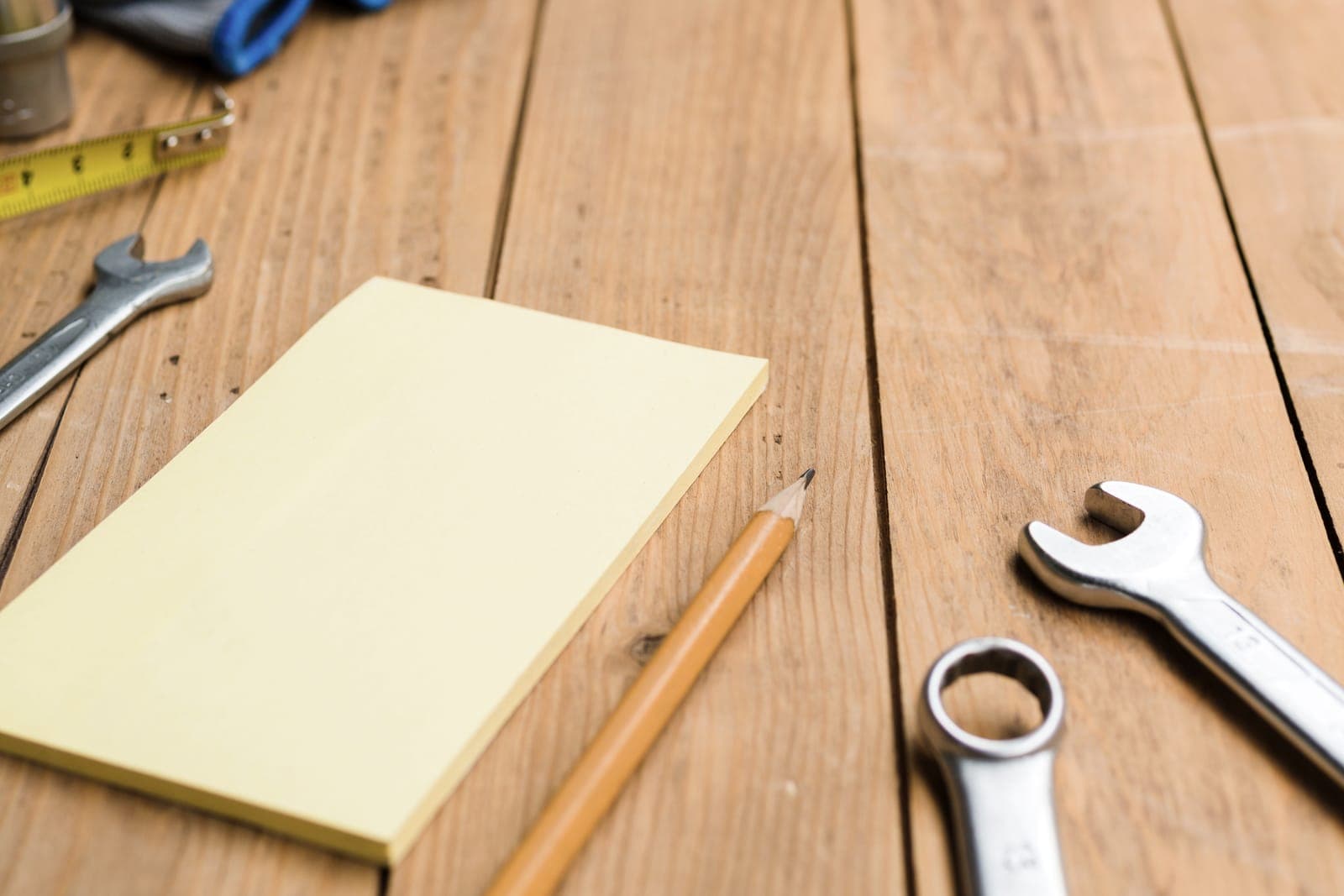 A canary-yellow notepad and a pencil on a plank workbench, wrenches and a tape measure around them.