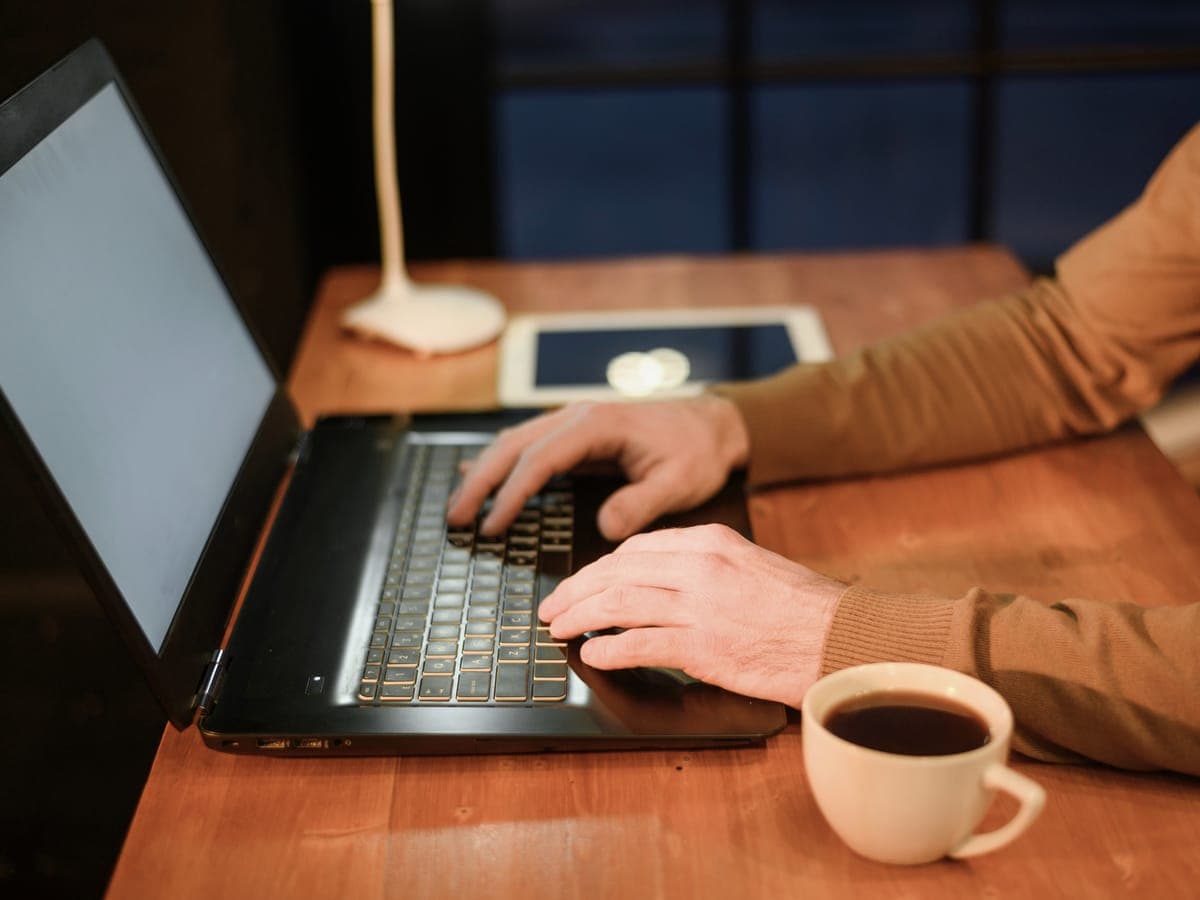 Hands typing on a laptop at a wooden table in the evening, a cup of coffee beside it.