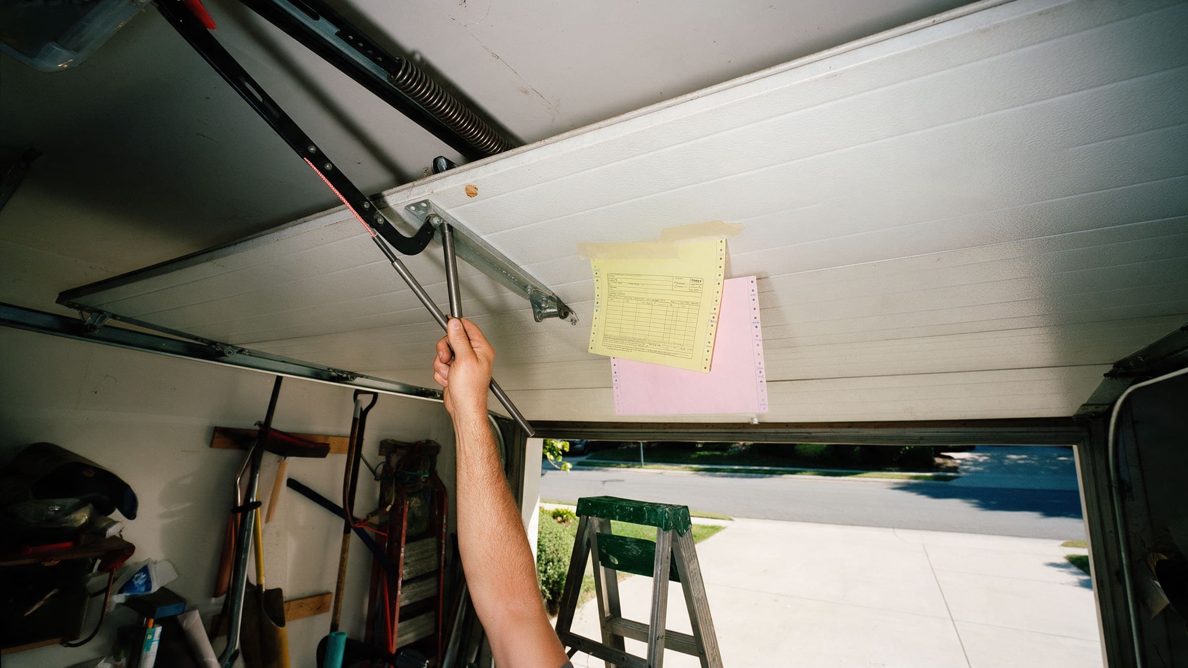 Inside a garage: an arm working on the torsion spring of a half-open door, carbon copies taped to a panel.