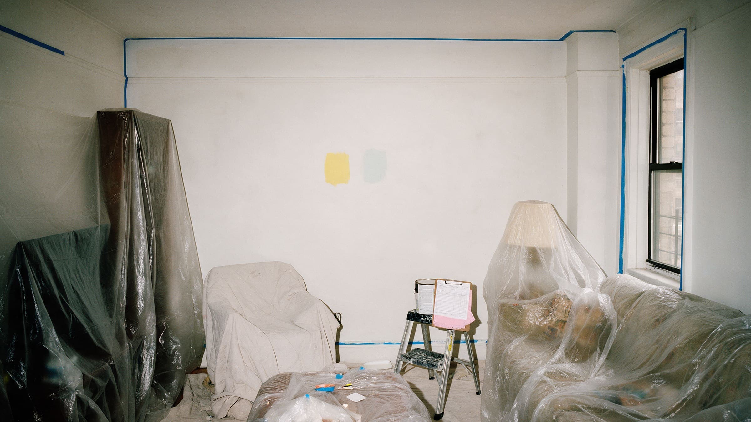 An empty living room prepped for painting: furniture under plastic, blue tape on the trim, two color swatches on the wall.