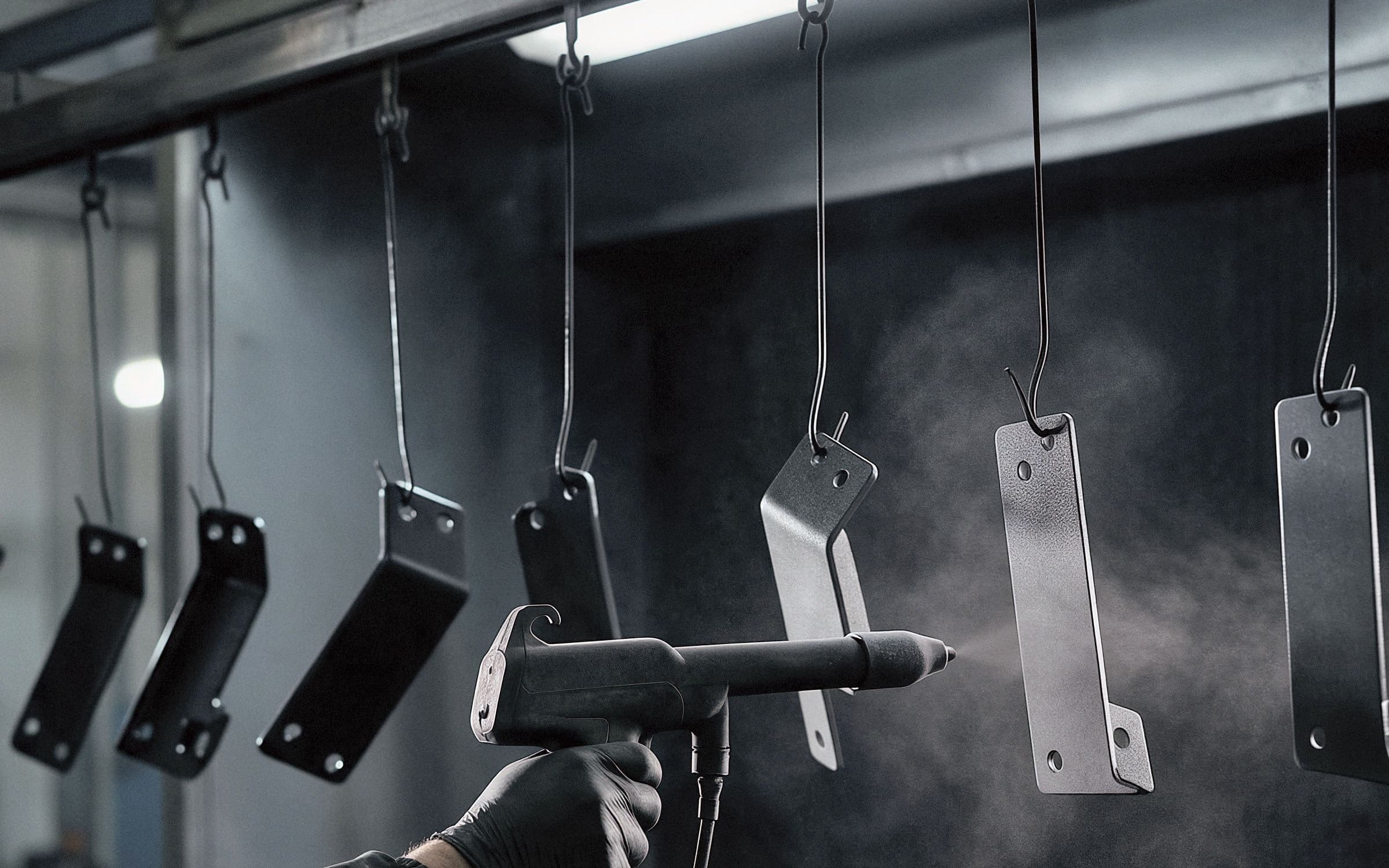 Gloved hand spraying black powder on brackets hanging from a rail in the booth