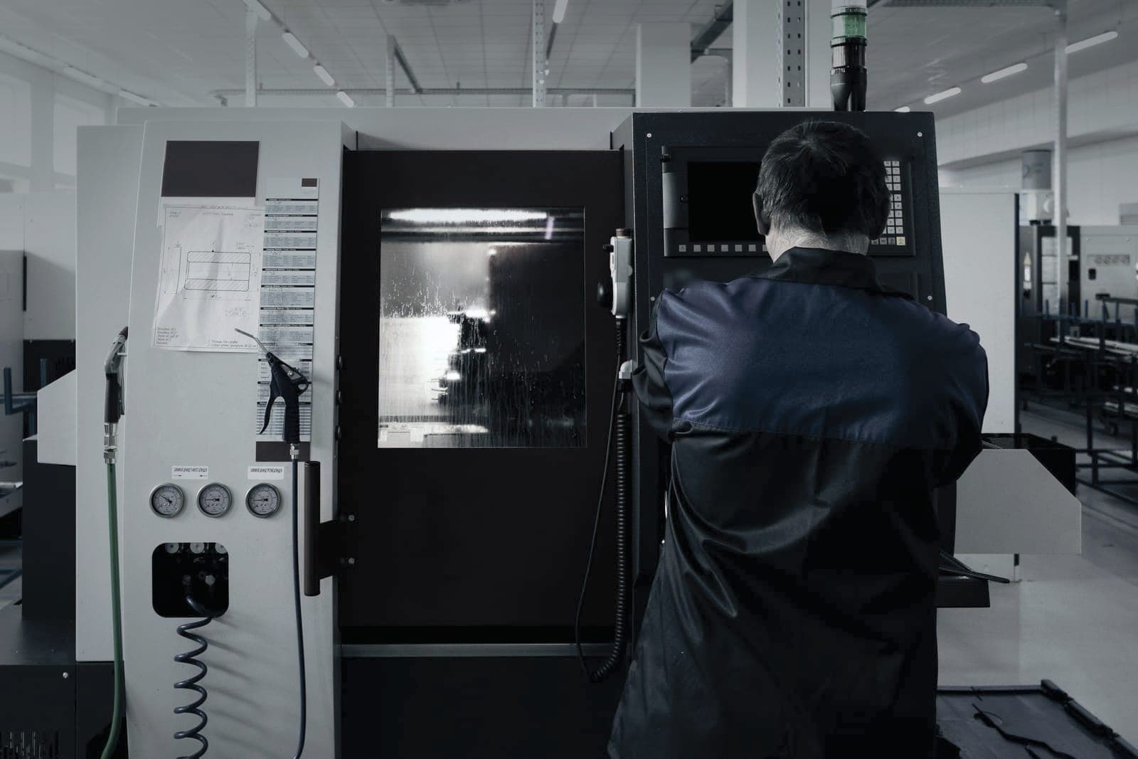 Machinist at the control of the CNC lathe, seen from behind, the part spinning behind the coolant-streaked window
