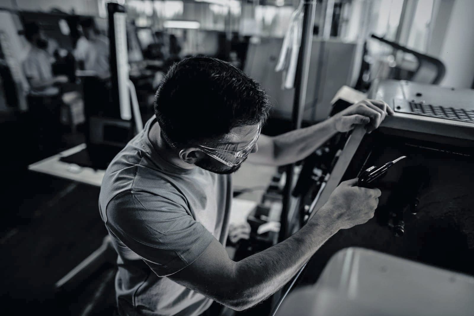 Machinist in safety glasses blowing chips out of a machine with an air gun, two colleagues talking behind him