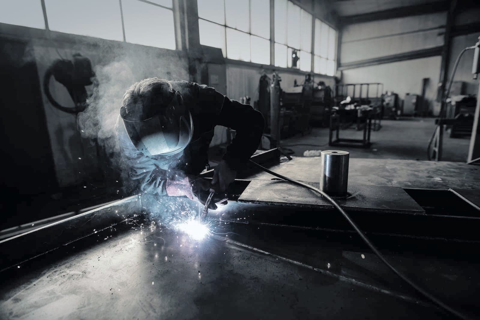 Welder in a helmet running a MIG bead on a large plate, smoke drifting toward the high windows