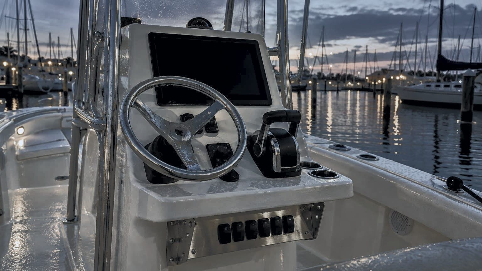A boat helm at dusk in the marina, a stainless bracket under the console holding the switch panel, harbor lights behind
