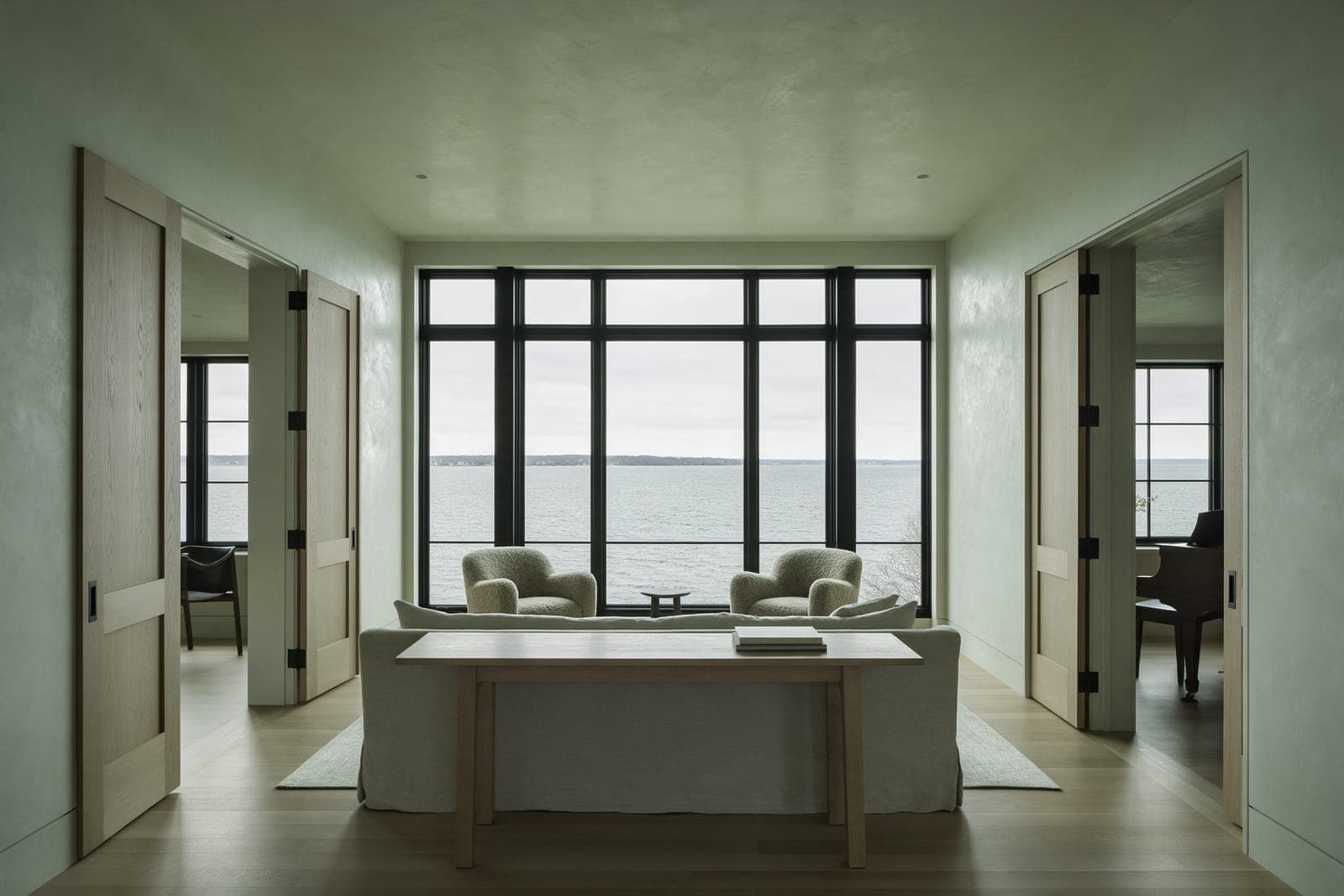 The living level of Bluff House, a long table and two armchairs in front of tall black windows on the Sound.