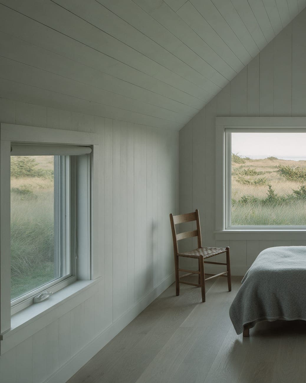 A corner of the east bedroom at Dune Shingle House: whitewashed board walls, a rush-seat chair and windows on two sides onto the dune grass.