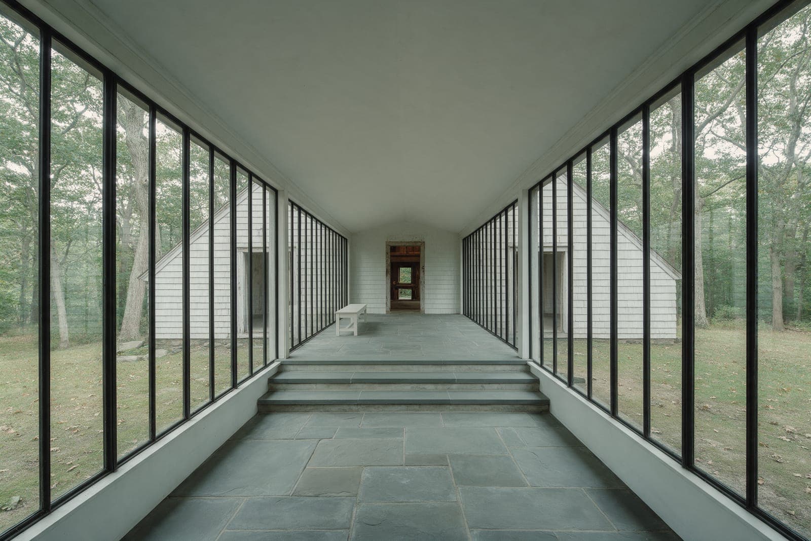 Inside the glass hall at Gable Pair: a bluestone floor stepping down between walls of glass to the old house.