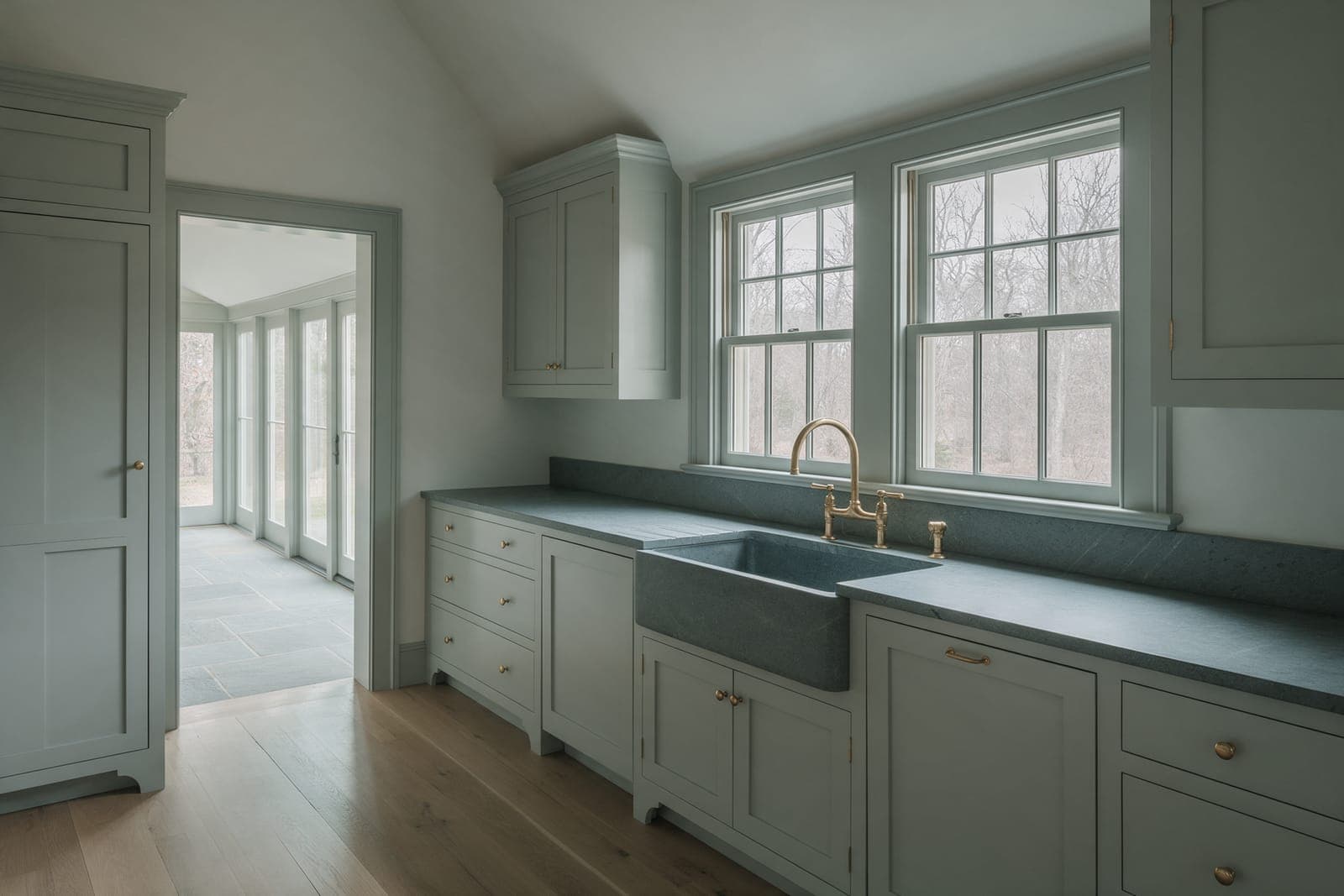 The one kitchen at Gable Pair: grey-green painted cabinets, a soapstone counter and farmhouse sink, the glass hall through the opening.