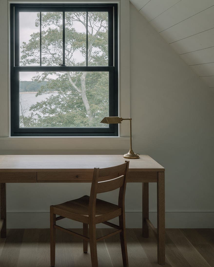 The study in the Harbor Hill wing: an oak desk and chair under a black double-hung window, a brass lamp, the harbor through the oak leaves.