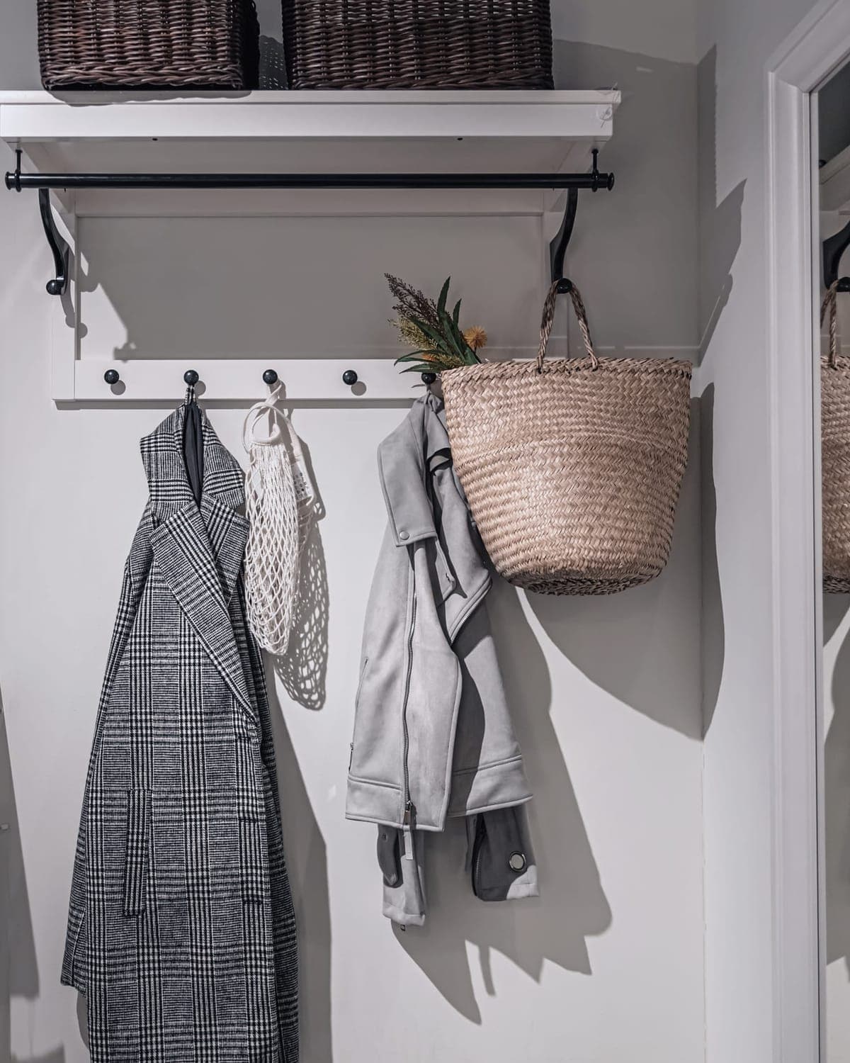 The mudroom wall at Harbor Hill: a white shelf with two wicker baskets, a black rail, a row of pegs with coats and a straw bag.