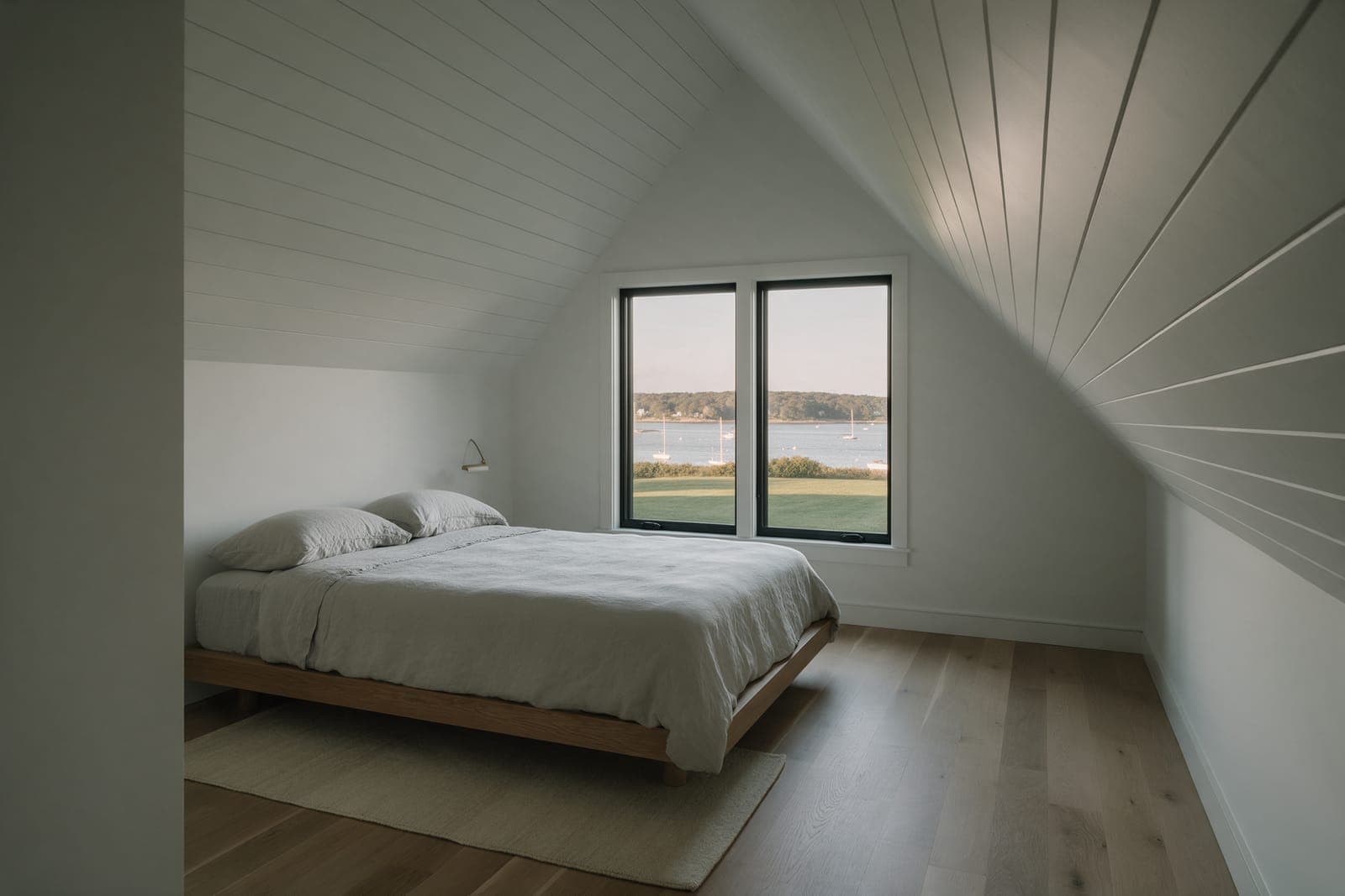 The primary bedroom in the Harbor Hill wing under a steep board ceiling, two windows looking down the lawn to the harbor.