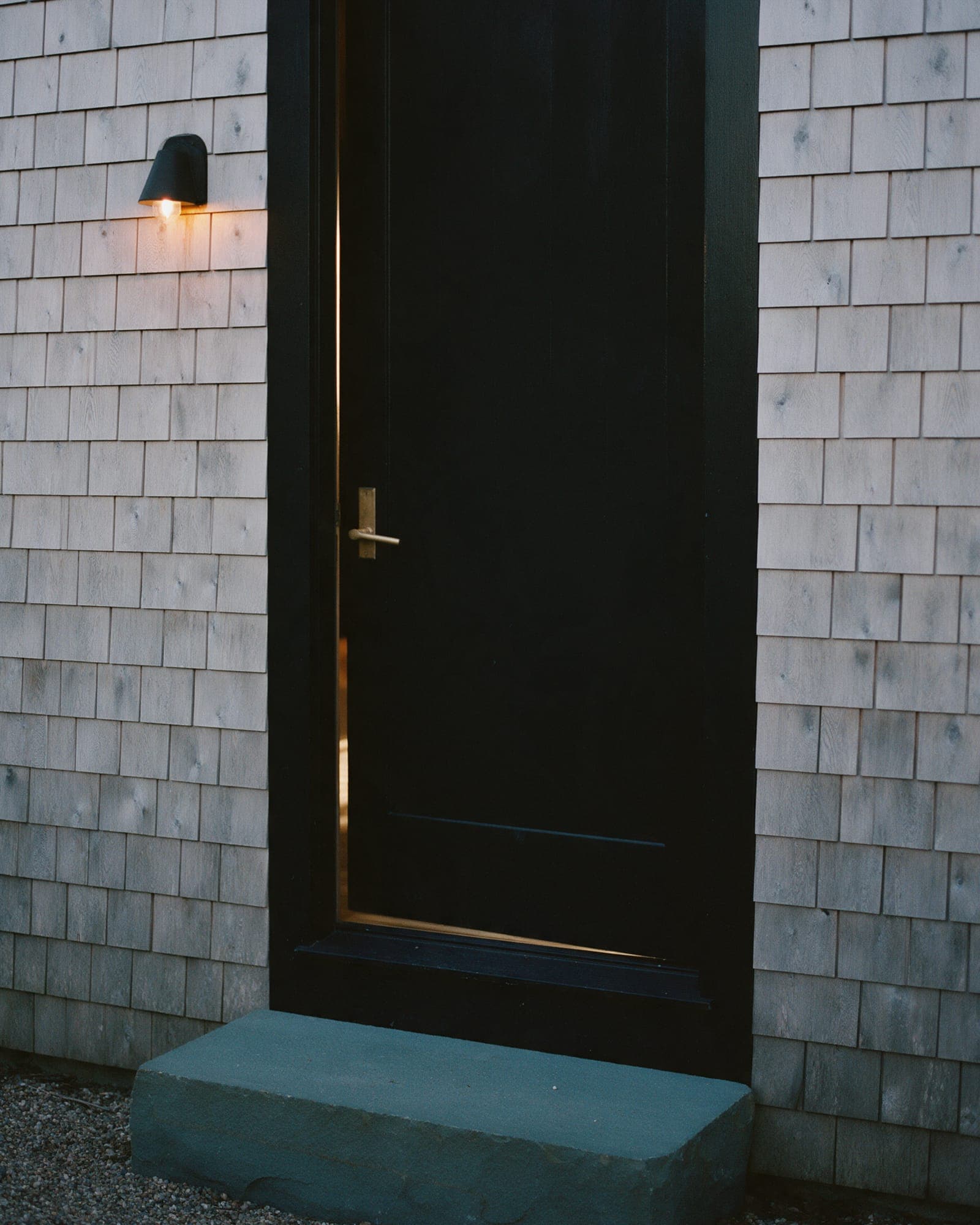 The studio door: a black-painted door in a silver shingle wall, a wall lantern lit above a wide bluestone step.