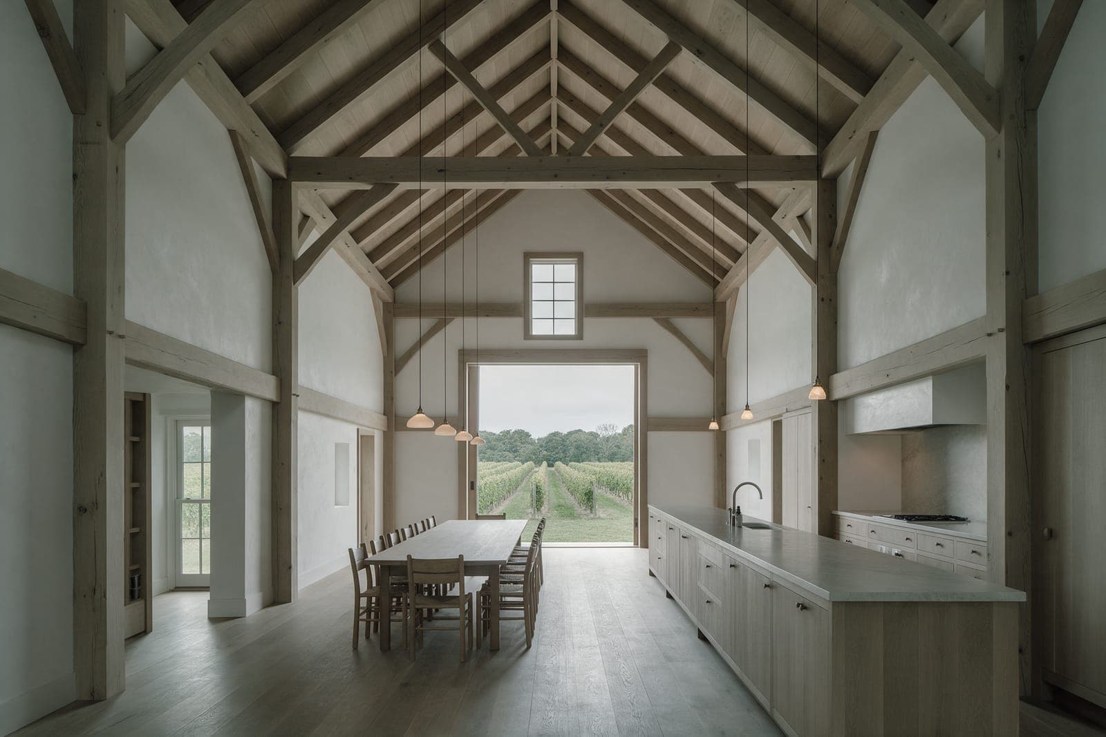 The double-height main room of Vineyard Barn House: timber frame, a long table and the sliding door open to the rows.
