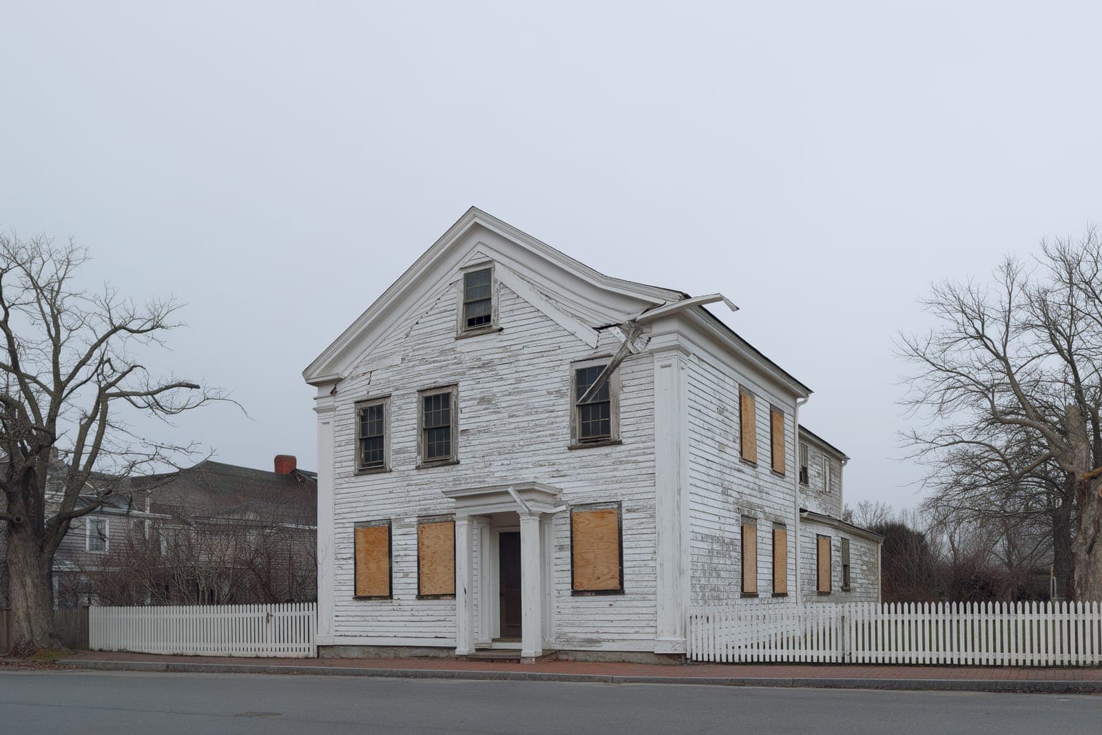 Whaler's Cottage before the work: peeling clapboard, plywood over two windows, a sagging porch roof and bare winter trees.
