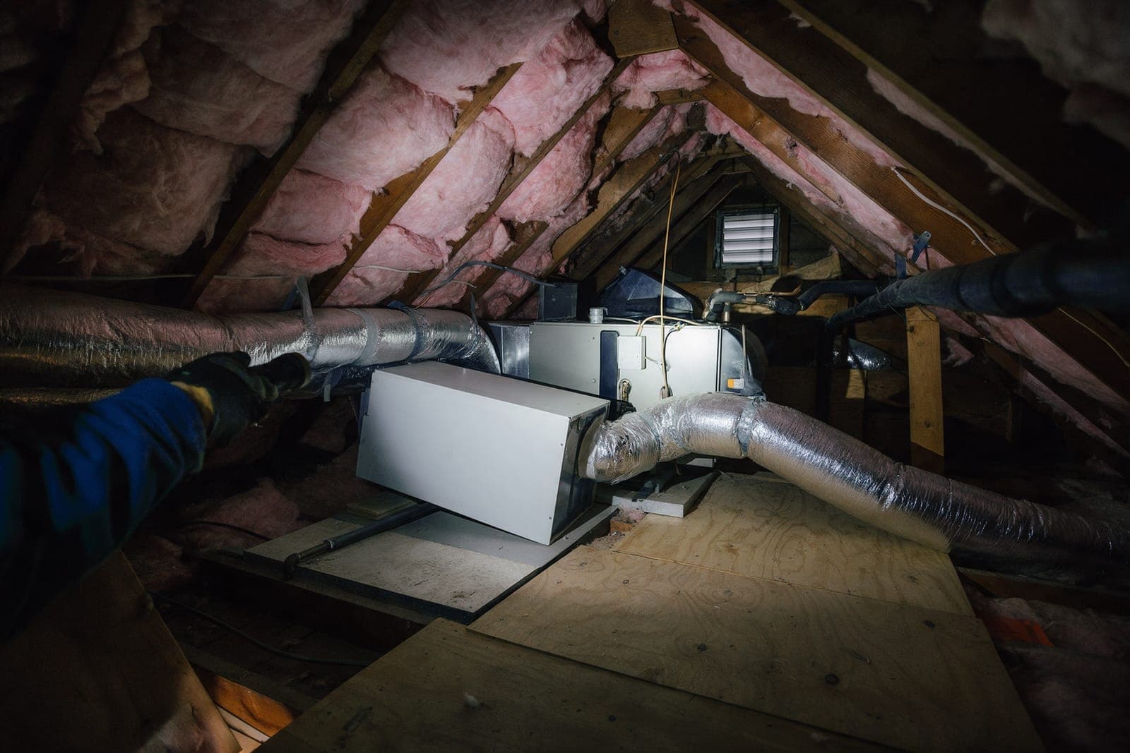 Compact air handler on a platform in a cape's knee-wall attic, lit by a technician's flashlight