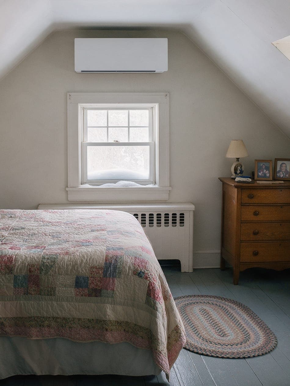 Upstairs cape bedroom under a sloped ceiling: a slim white ductless head above the window, snow on the sill, a patchwork quilt and an oak dresser