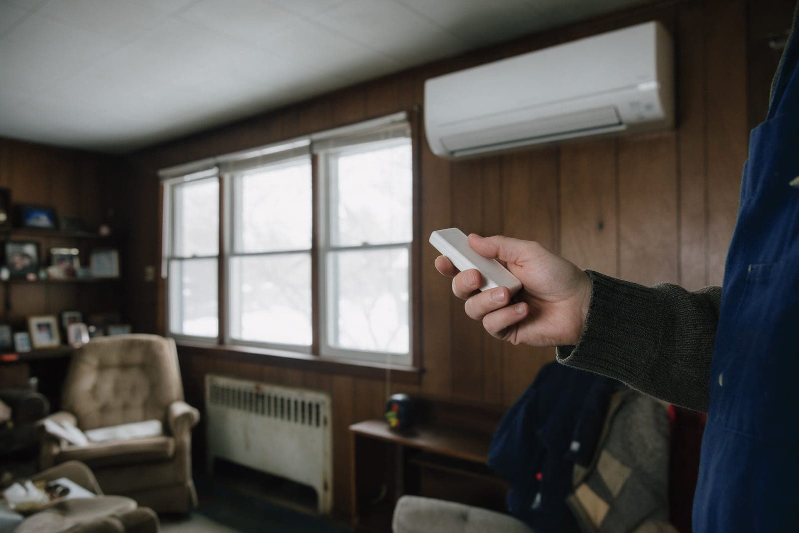 Ductless head mounted high in a wood-paneled 1960s living room, a homeowner's hand holding the remote