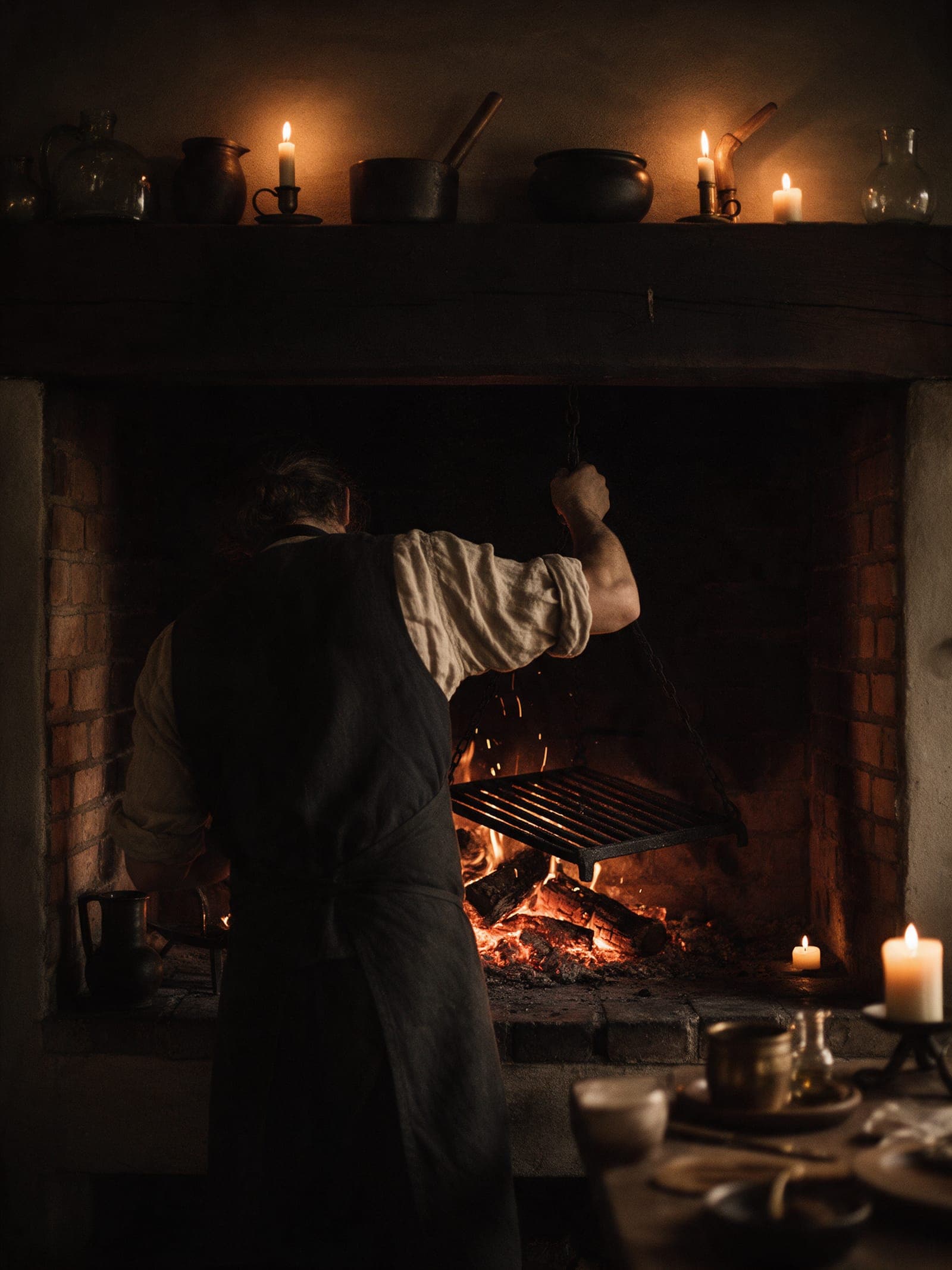 A cook seen from behind at the open hearth, raising the cast-iron grate on its chain over glowing oak embers.