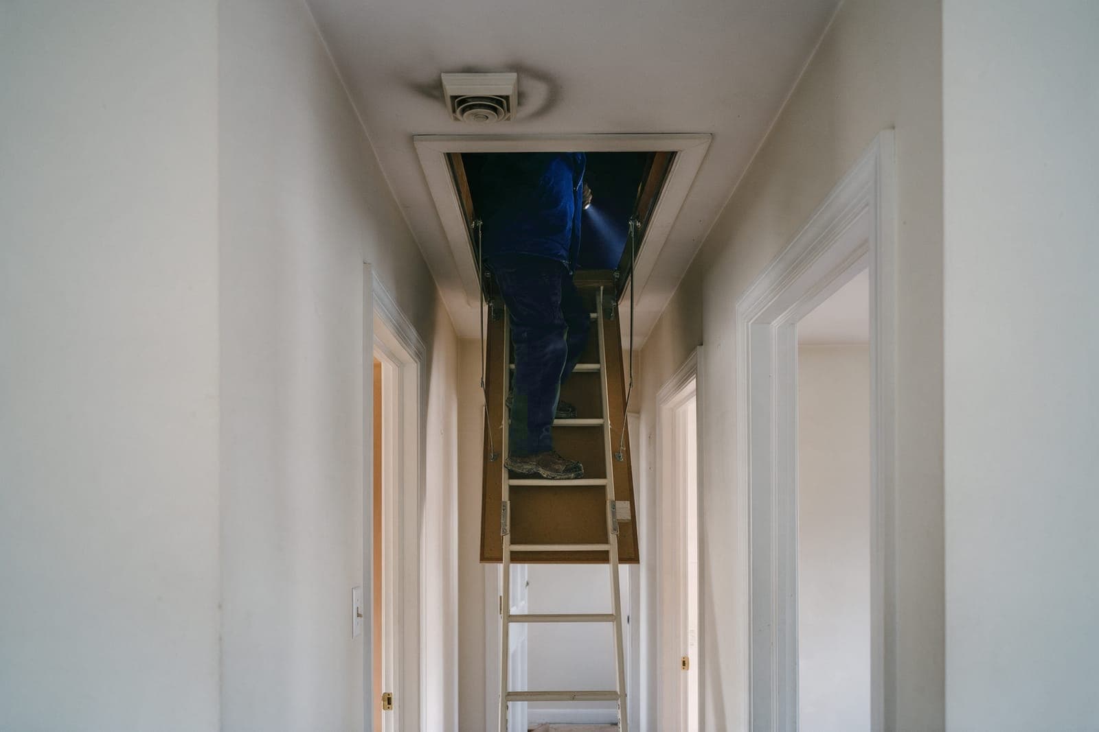 An upstairs hallway in a colonial, the attic ladder pulled down and a technician climbing into the attic with a flashlight, a dusty ceiling supply register beside the hatch