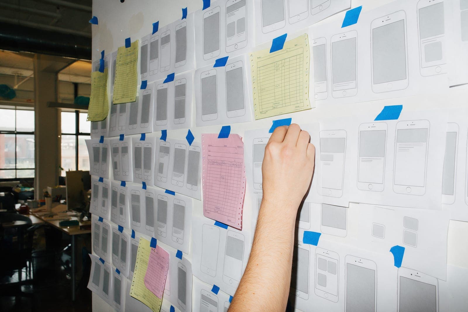 A wall of paper phone-screen sketches pinned with blue tape, canary and pink forms among them, a hand pinning one more.