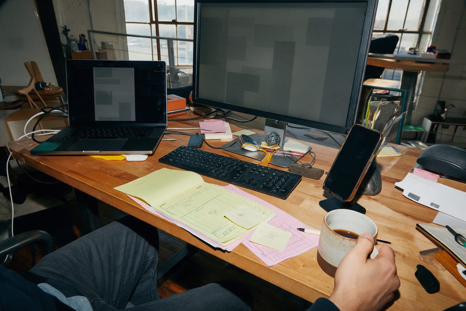 A loft office desk with a laptop, a monitor of grey blocks, a phone on a stand and a carbon estimate pad by the keyboard.