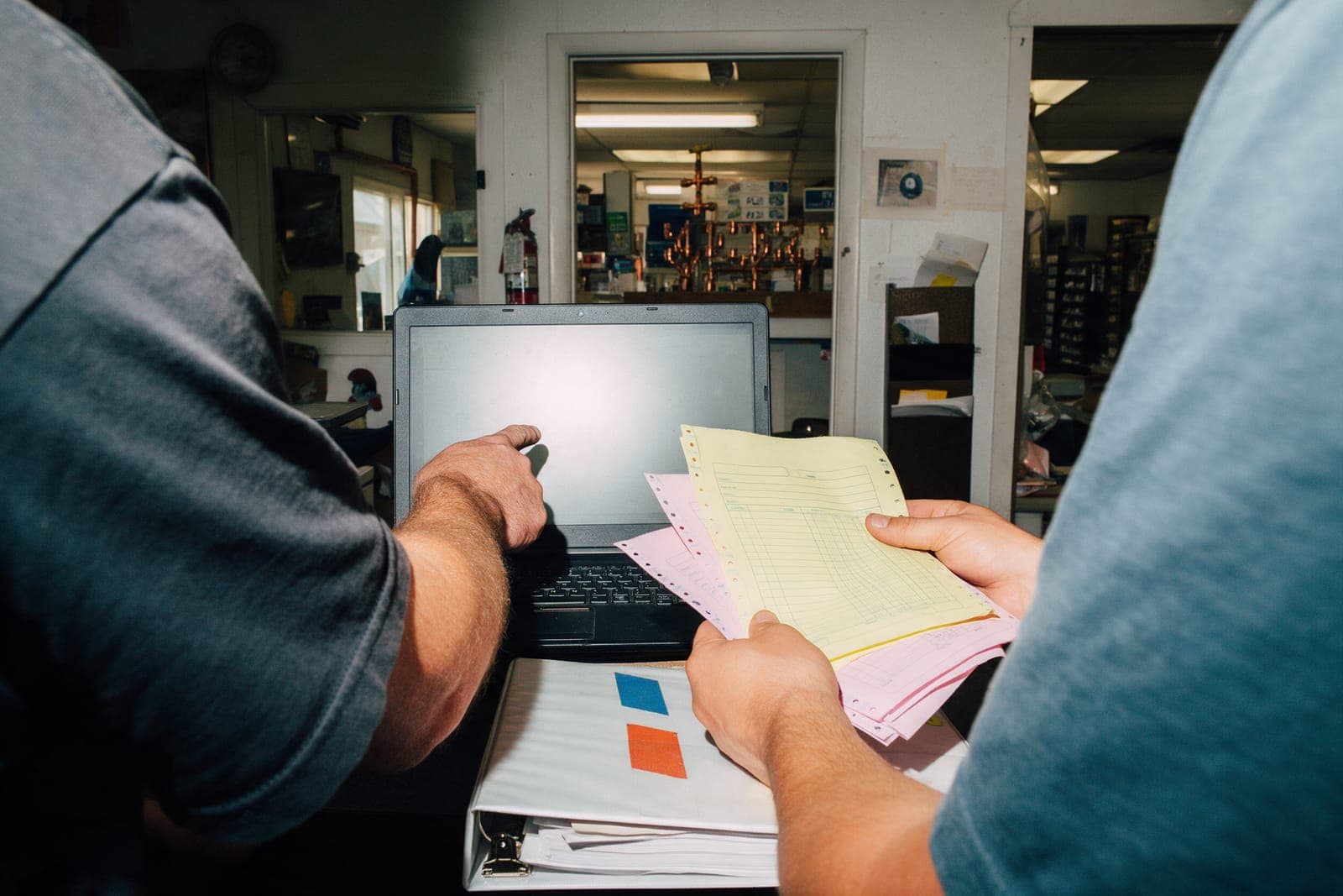 Two people at a plumbing shop desk, one pointing at a laptop, the other holding canary and pink copies, a parts counter behind.