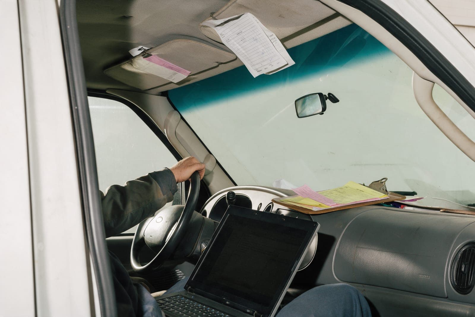 A ride-along in a work van: a driver's arm on the wheel and a laptop on the passenger's lap.