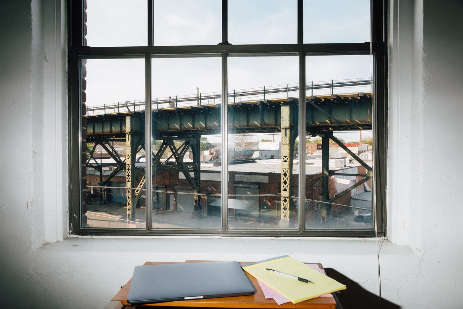 A loft window looking out at an overhead train line and warehouses, a yellow pad on the desk.