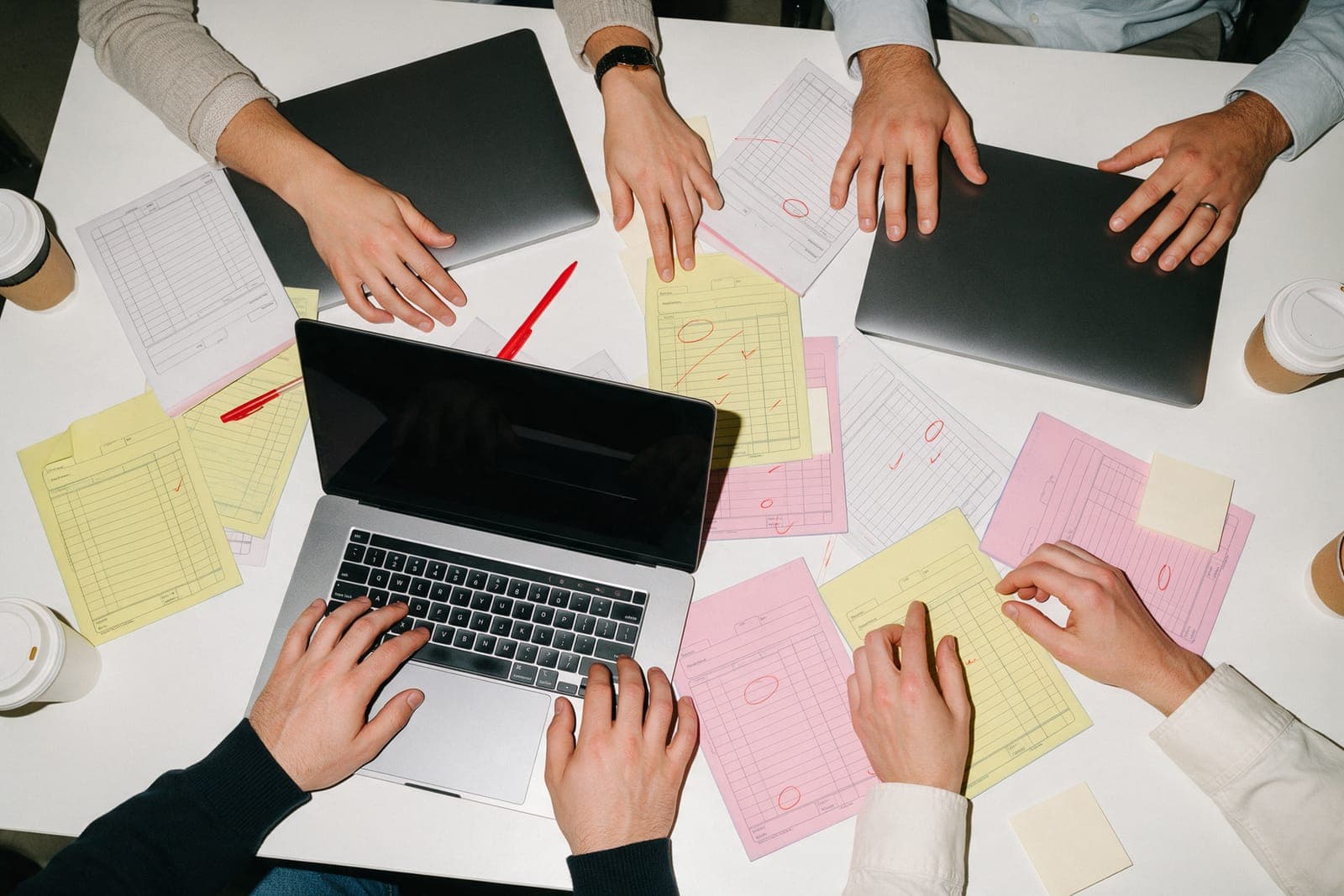 Four people's hands around a table of laptops and printed quotes marked in red.