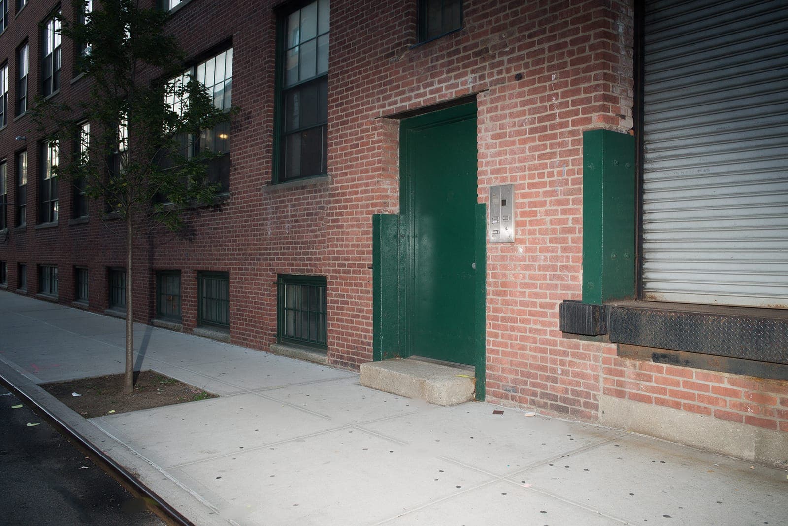A dark green steel door in an old brick loft building, a blank intercom plate beside it and a loading dock with a closed shutter to the right.