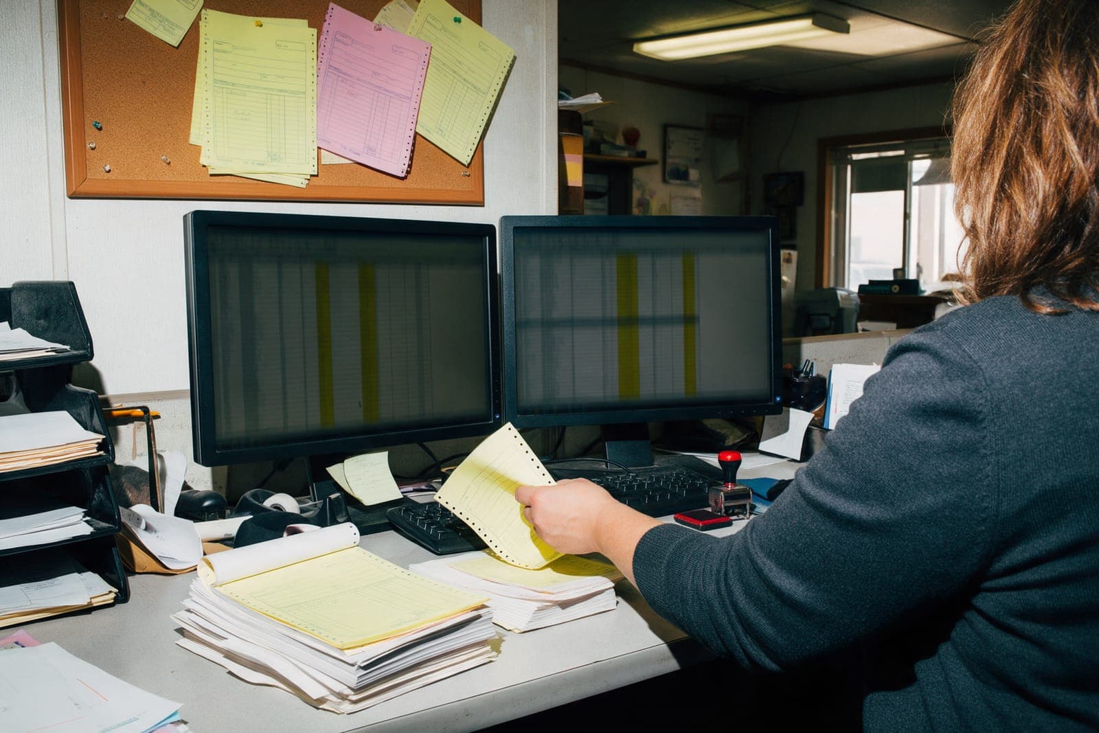 An office manager from behind at two monitors, moving a canary copy between stacks under a corkboard of pinned copies.