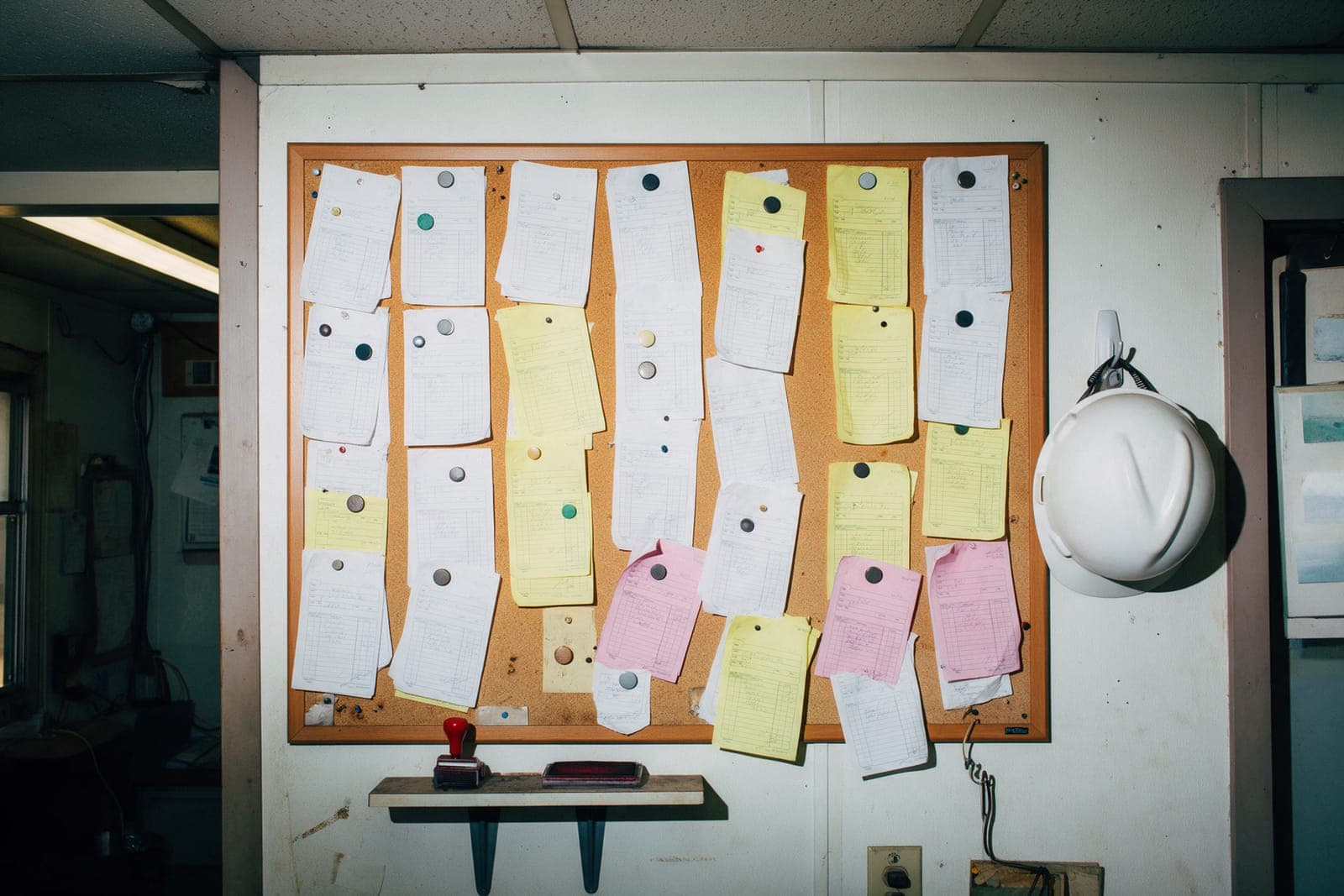A corkboard of pinned carbon copies in rough columns, a rubber stamp on the shelf below.